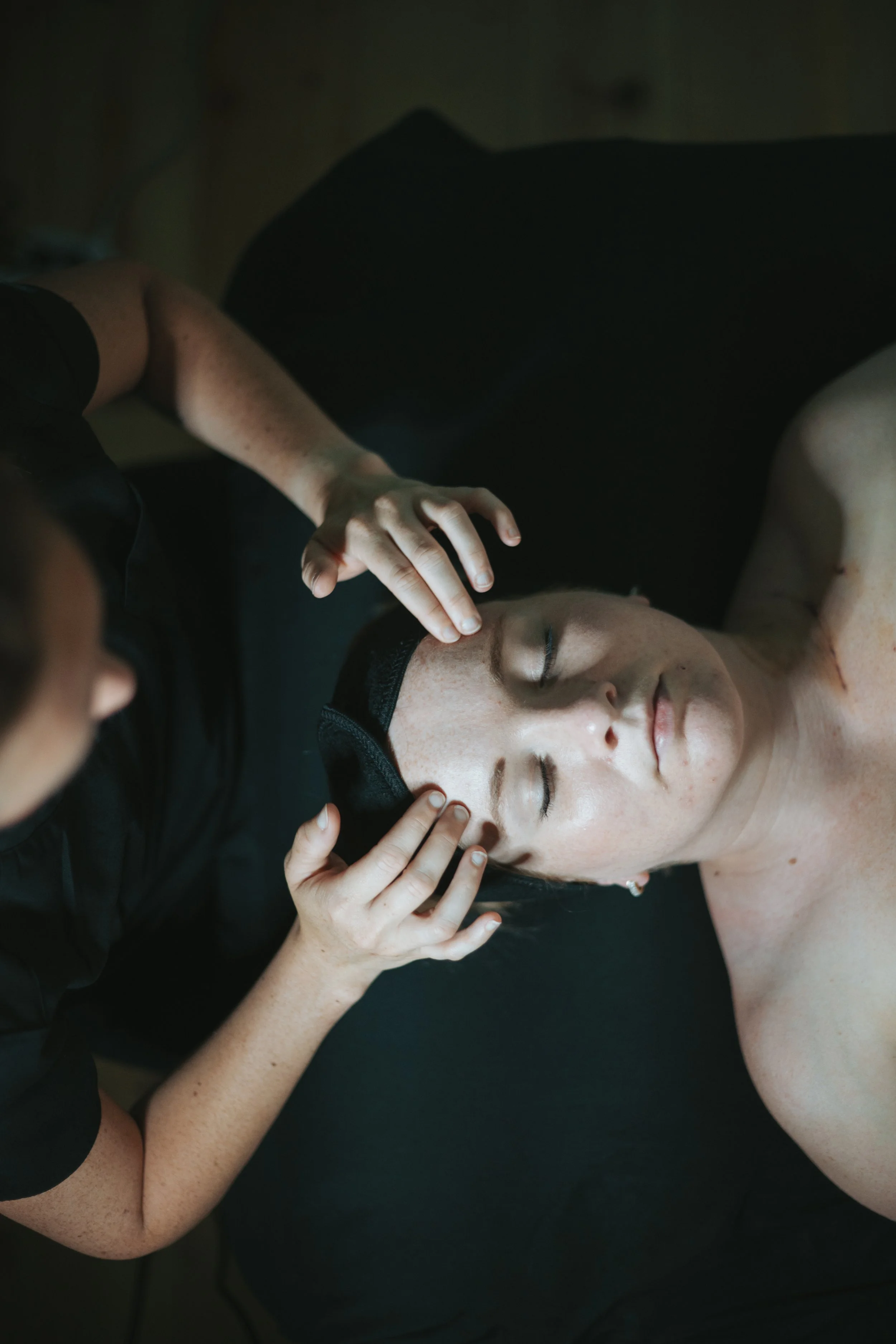 A woman with closed eyes receiving a facial massage from a massage therapist in a dark room.