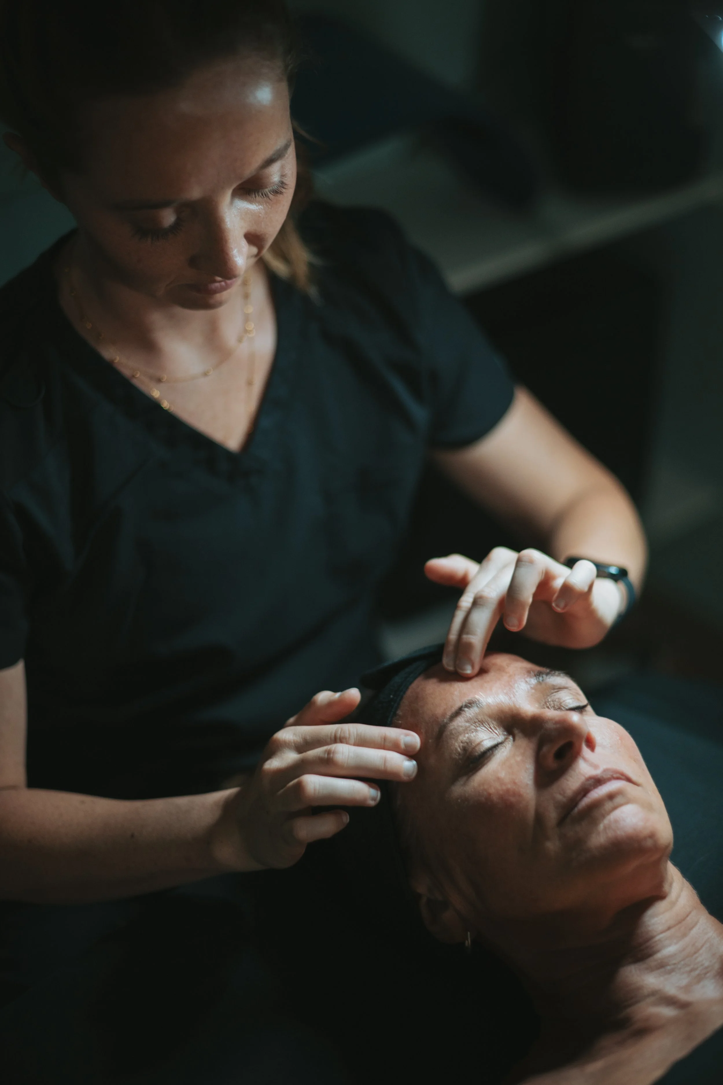 A woman with freckles and red hair practicing facial acupressure or massage on a middle-aged woman with closed eyes, relaxed expression, and dark hair, while lying down.