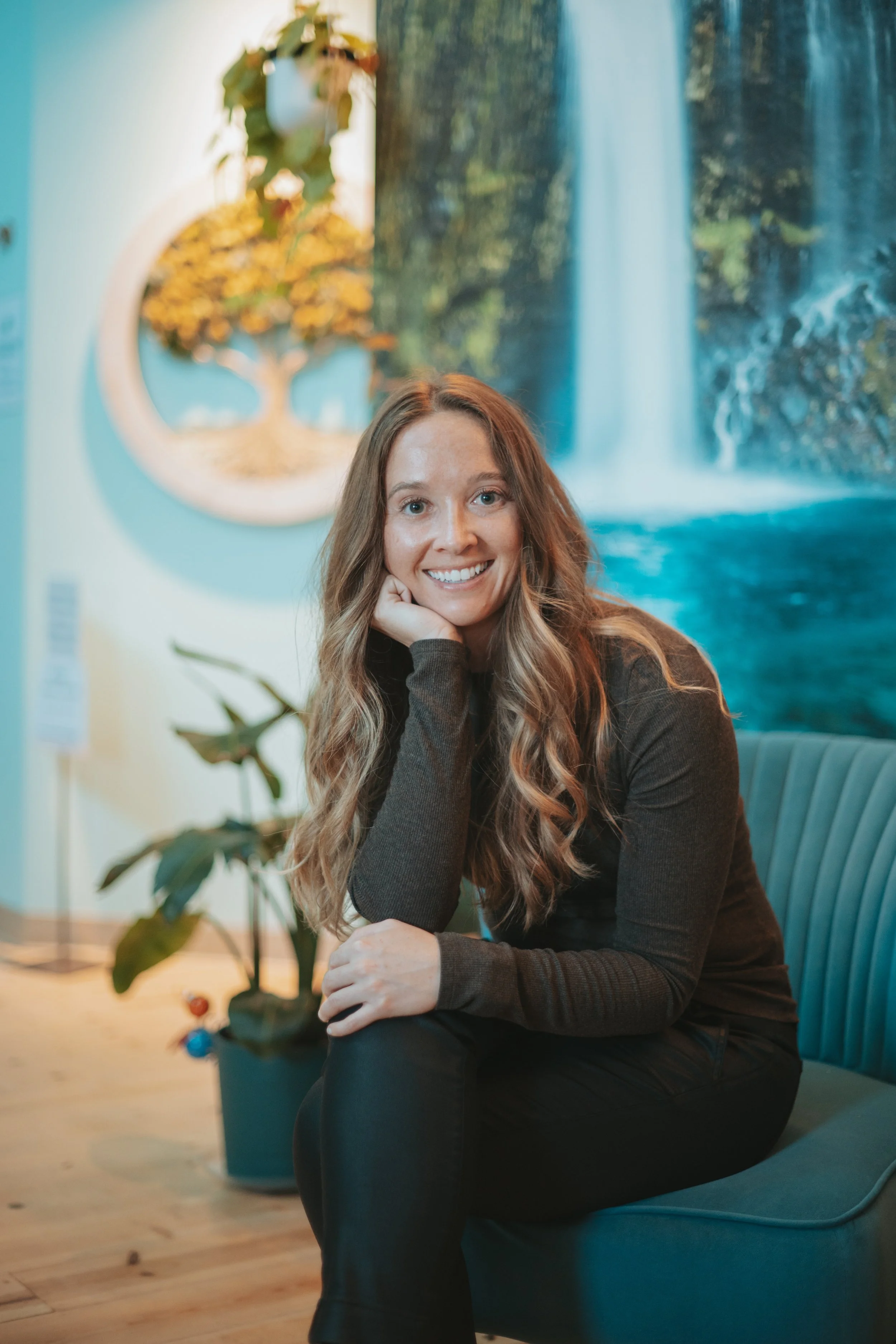 A woman with long wavy hair, smiling, sitting on a blue chair, resting her chin on her hand, in a cozy indoor space with abstract blue waterfall wall art behind her. A potted plant with green leaves is partially visible in the foreground.