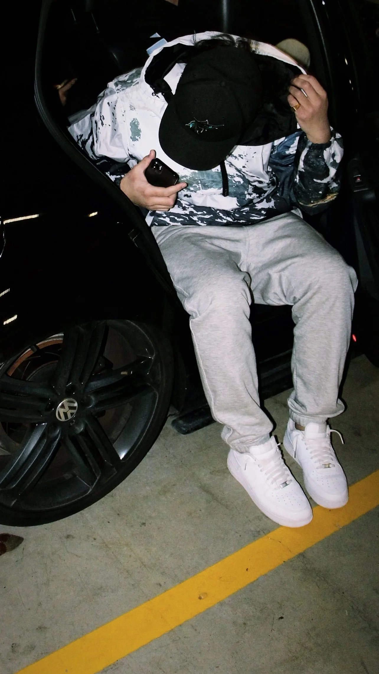 Person sitting in a wheelchair, wearing a white and black jacket, gray sweatpants, and white sneakers, holding a phone, with a black hat covering their face.