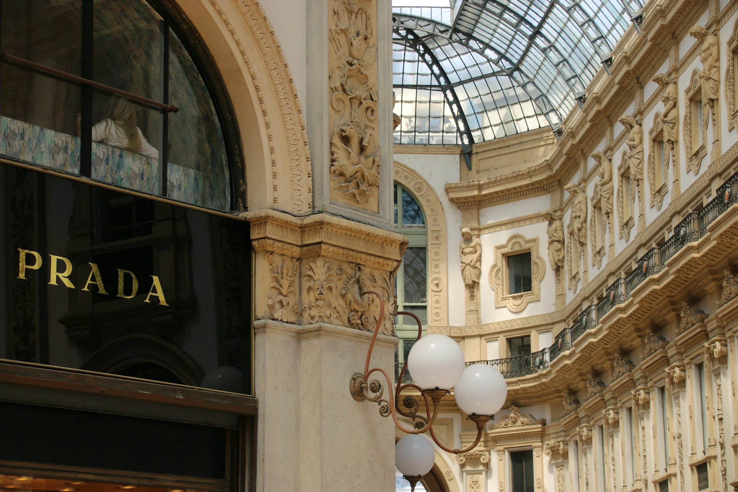 View of an ornate shopping gallery with detailed stone carvings, decorative sculptures, and a glass ceiling. A Prada store is visible at the bottom left.