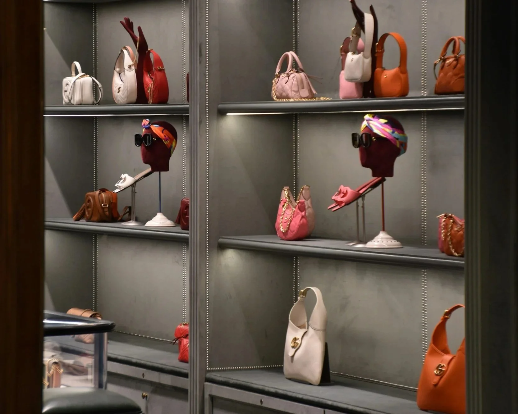 Display of various handbags on glass shelves, including white, red, pink, orange, and brown bags, with some accessories like sunglasses and headscarves on mannequin heads.