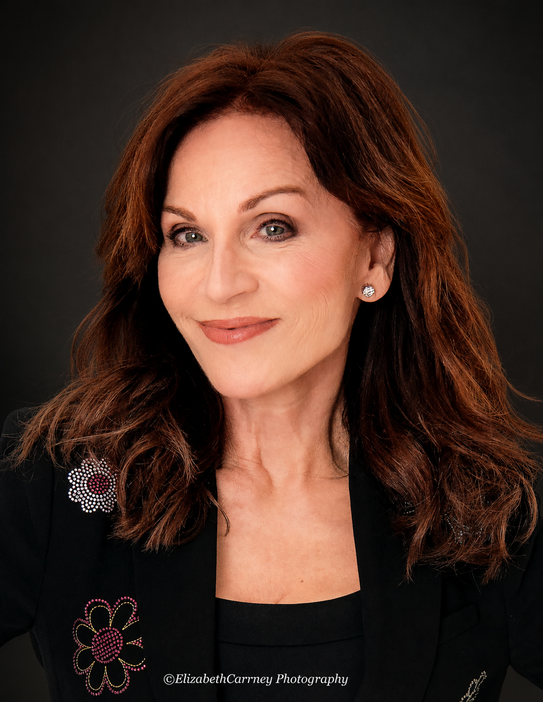 A woman with red hair wearing a black jacket with floral embellishments, smiling against a dark background.