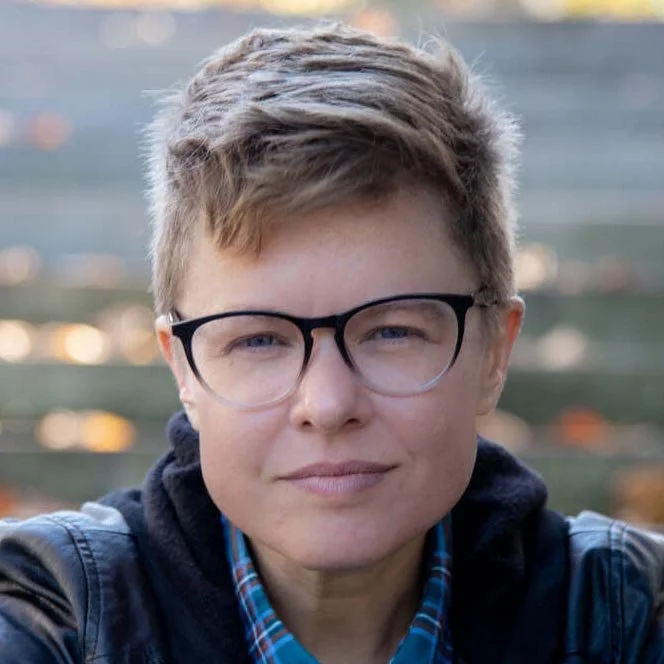 Close-up of a person with short light brown hair, wearing black-rimmed glasses, a dark jacket, and a plaid shirt, outdoors with a blurred background.