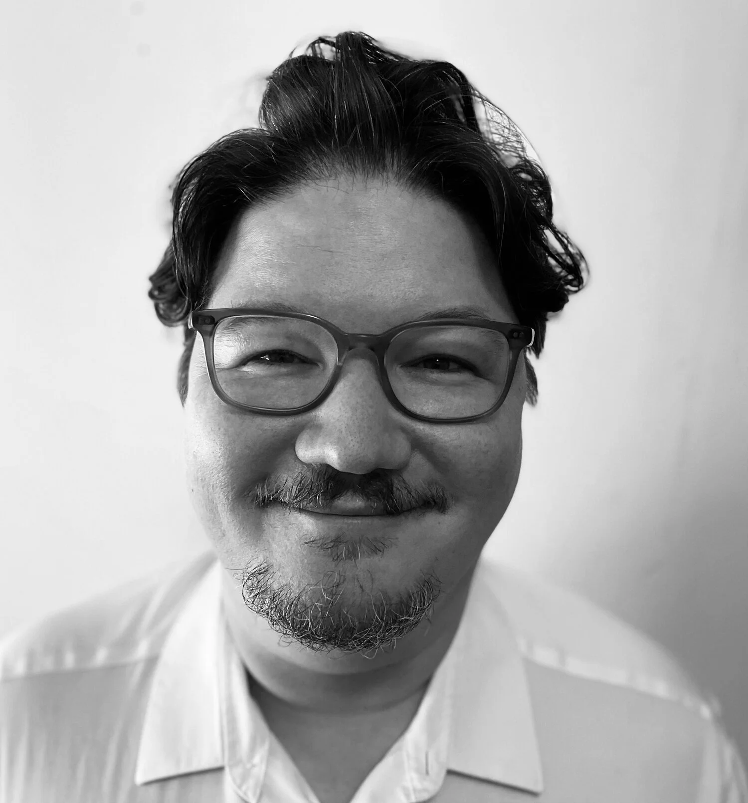 Black and white photo of a smiling man with glasses, short wavy hair, and a beard, wearing a collared shirt.