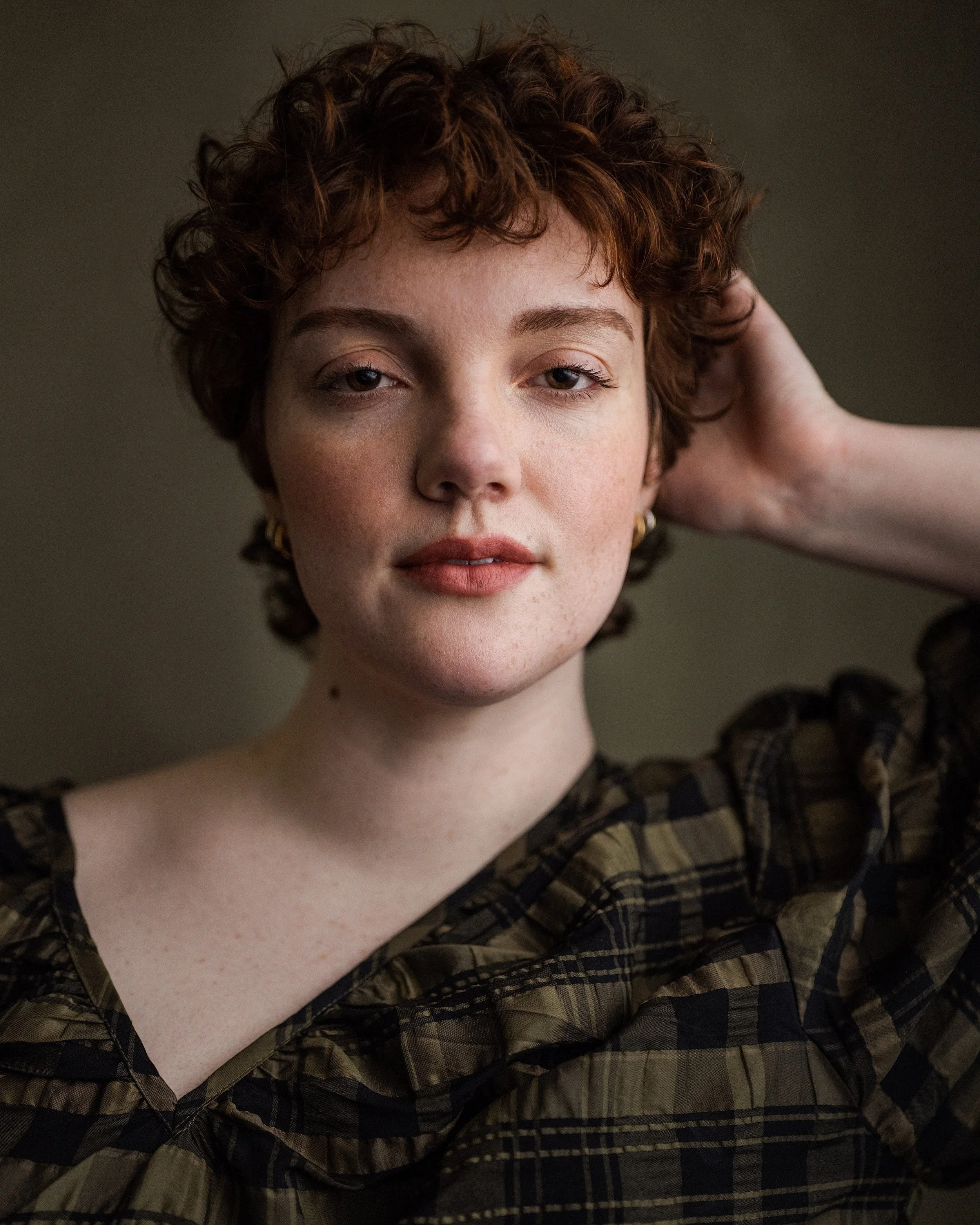 Close-up of a young woman with short curly brown hair, wearing gold hoop earrings and a plaid shirt, resting her hand on her head.