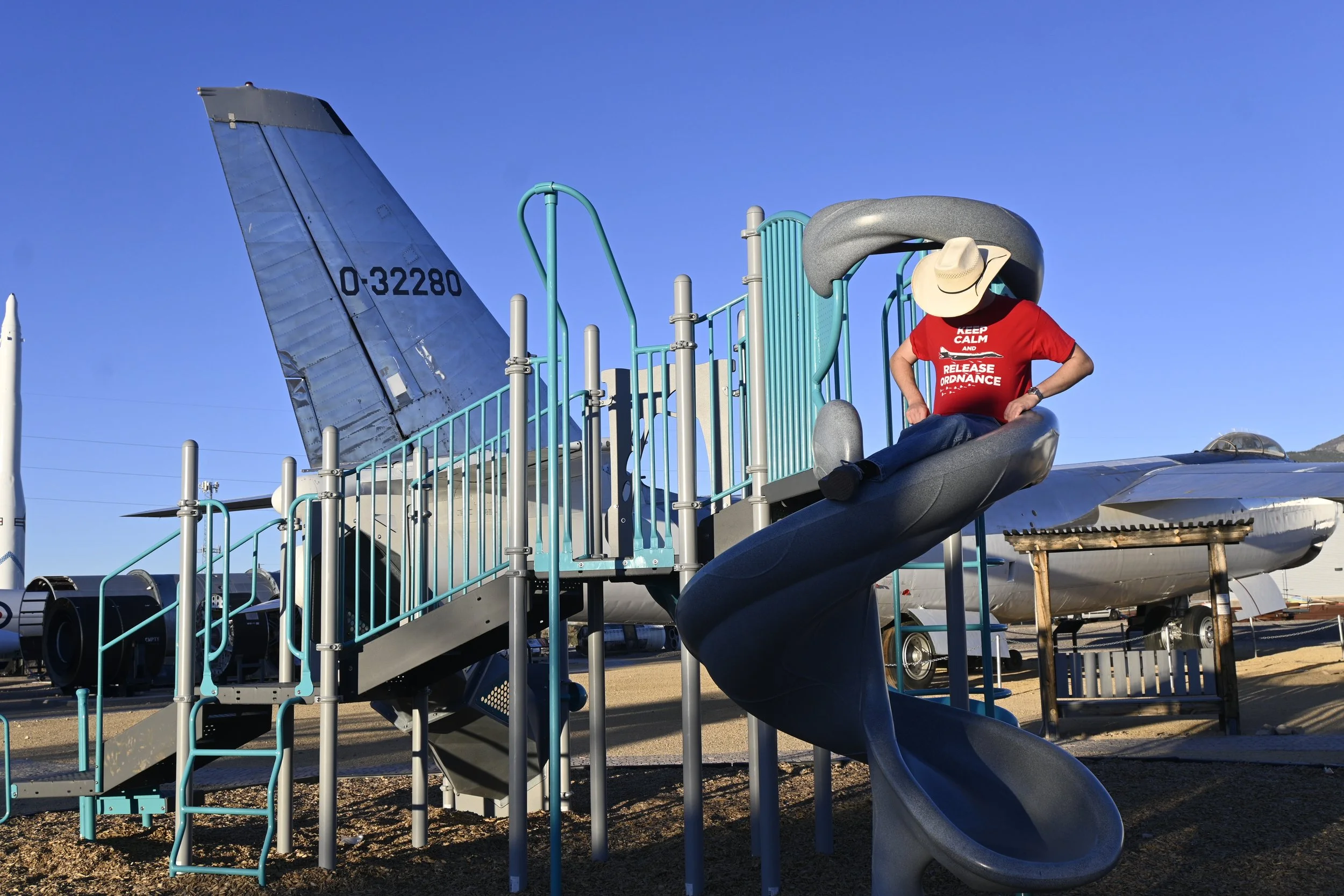Person sitting on playground slide wearing a cowboy hat and red shirt with white text, in an airplane graveyard with aircraft parts in the background.