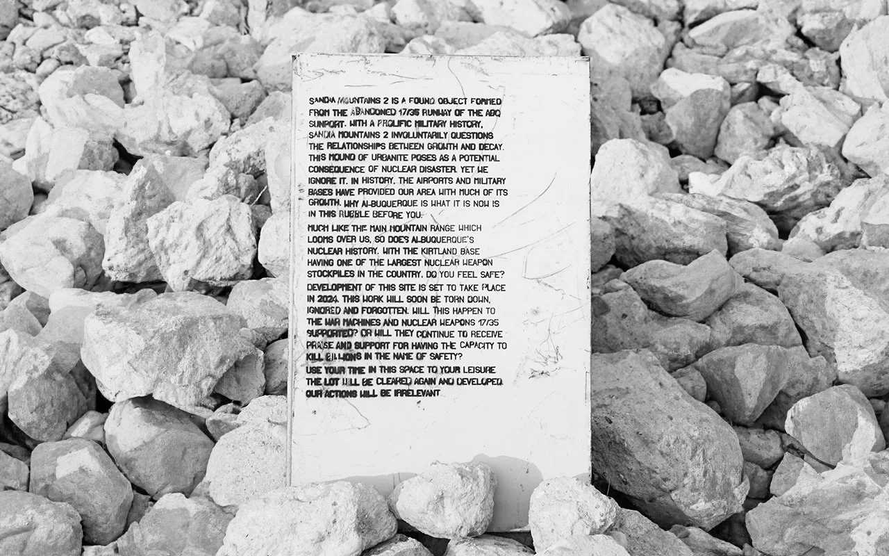 A printed sign on a white paper placed among rocks and gravel, with visible cracks and text discussing Sandia Mountains and nuclear history.