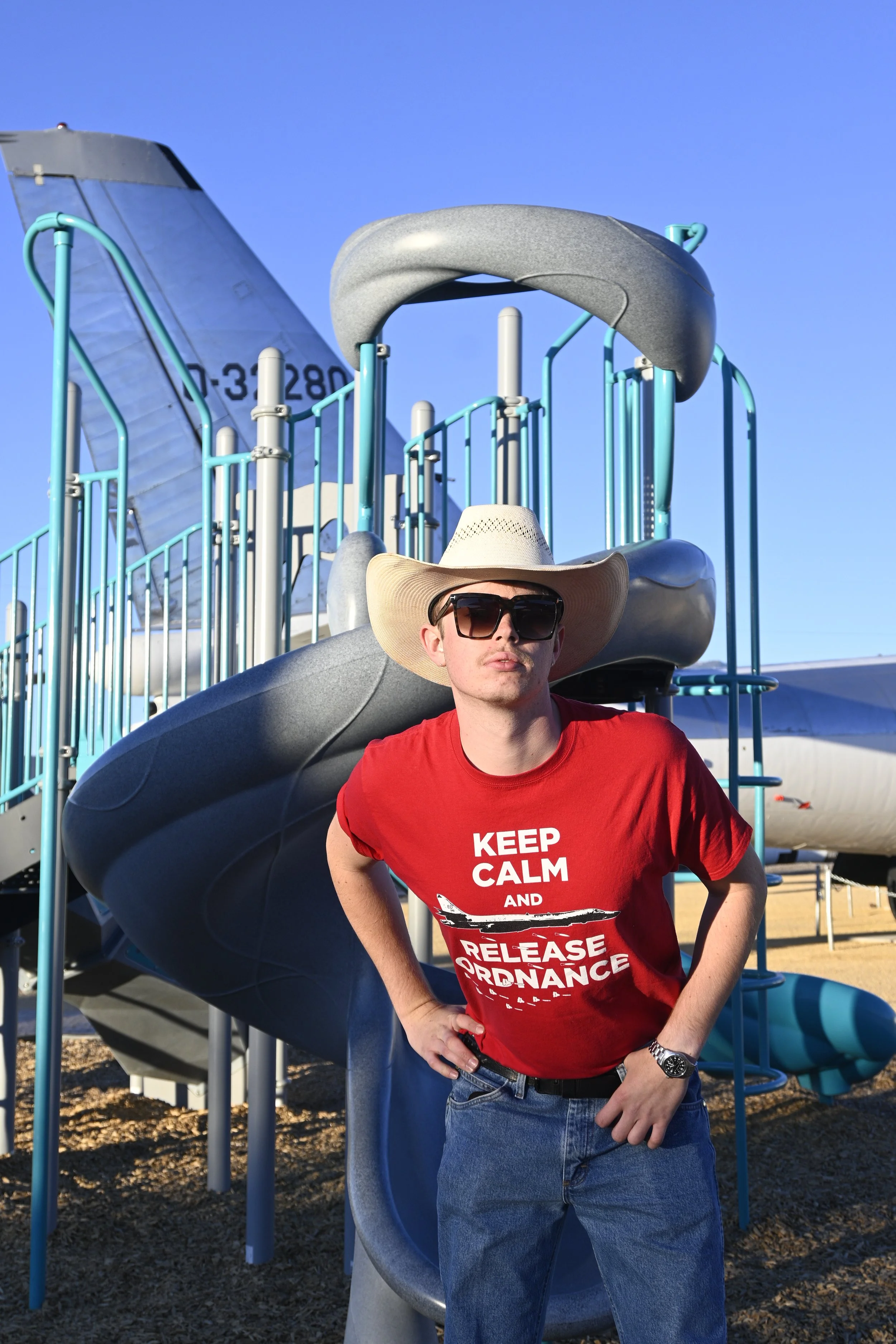 A man wearing a wide-brimmed hat and sunglasses, standing in front of a playground slide at a park on a bright day. He is wearing a red T-shirt with white text.