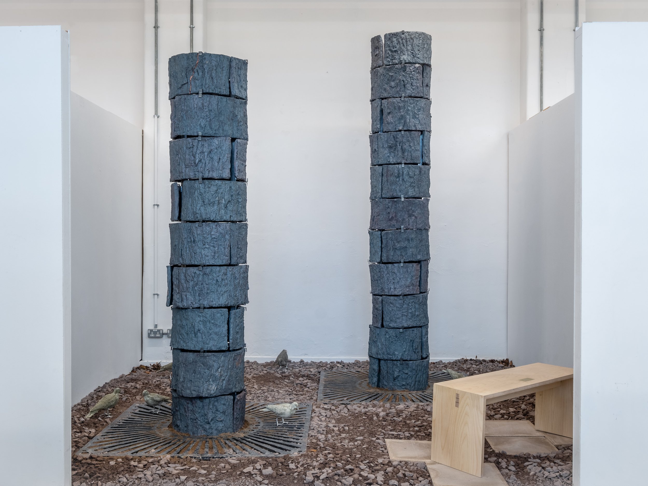Two tall columns made of roughly stacked dark gray stones inside a white-walled room with exposed pipes. Small birds are on the ground among gravel, and a wooden bench is positioned to the right.