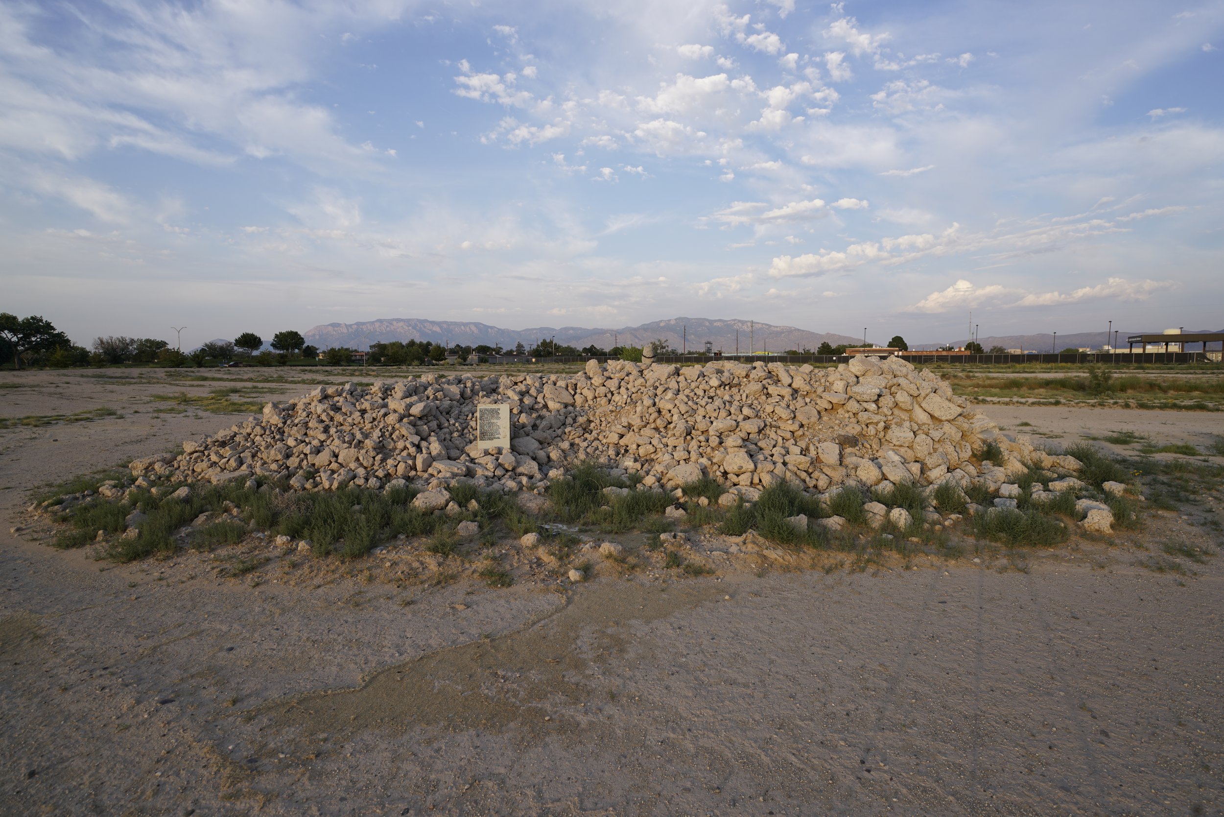 A pile of rocks in a barren area with some greenery, with mountains in the distance, a parking lot, and buildings under a partly cloudy sky.