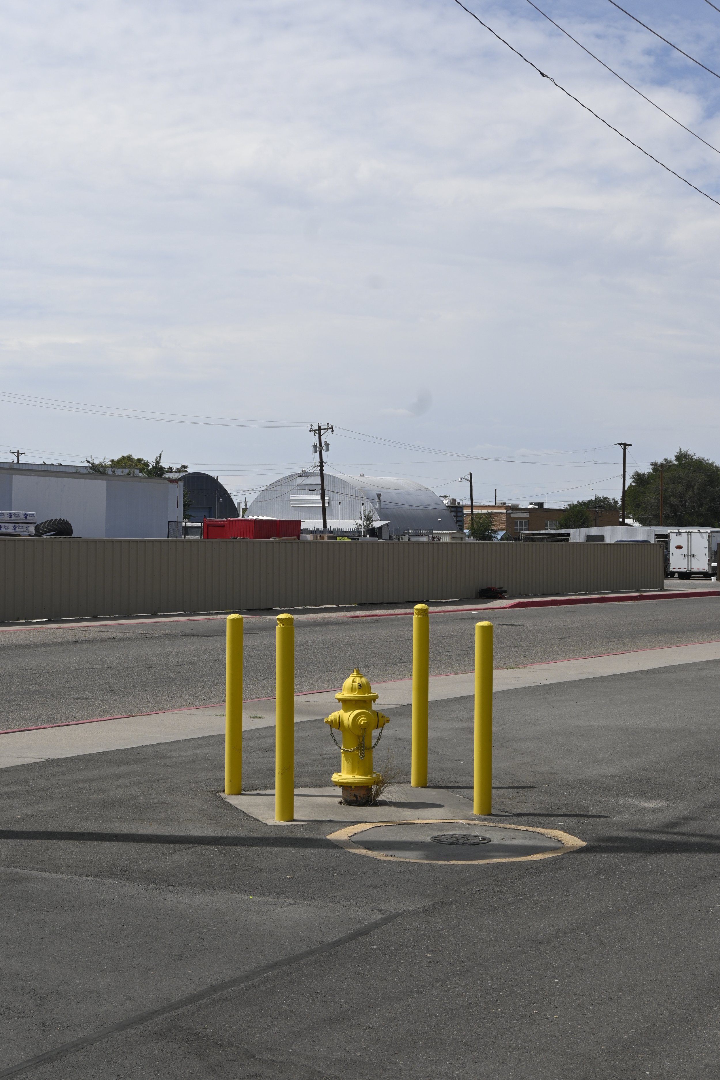 A yellow fire hydrant surrounded by three yellow poles on a paved sidewalk. Behind, there is a parking lot with a few vehicles, a fence, and industrial buildings with a cloudy sky overhead.