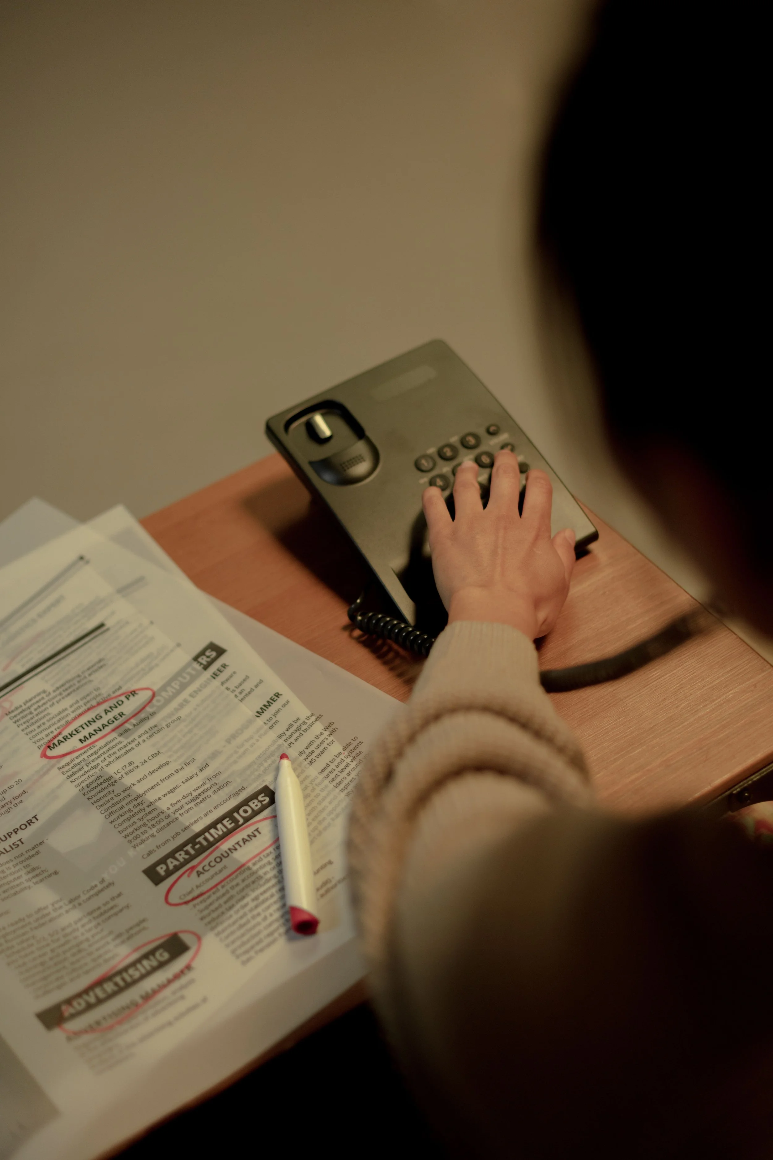 A person with a beige sweater uses a gray landline telephone on a wooden desk. On the desk, there are papers and a white pen with a red cap.