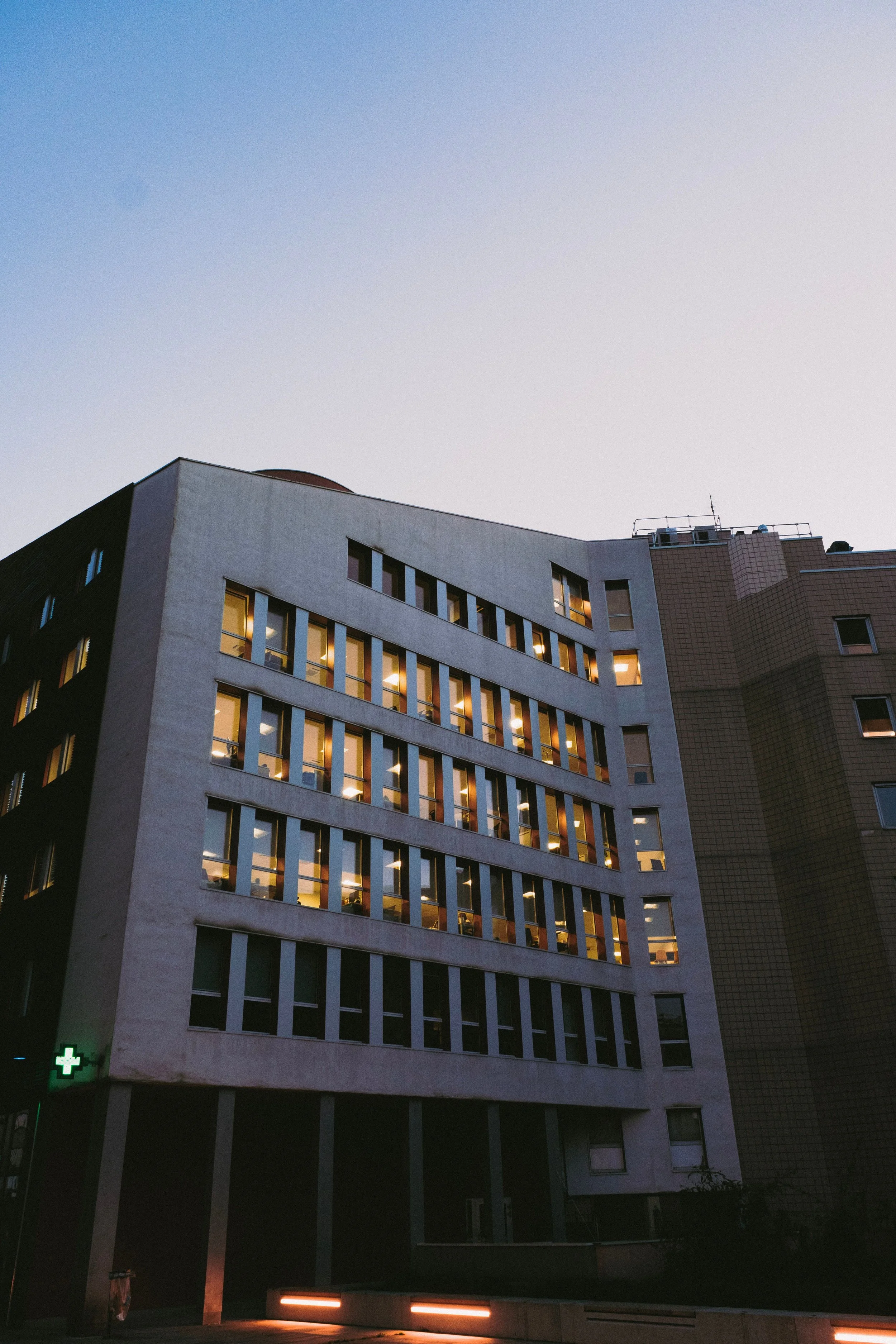 The image shows a modern multi-story building at dusk with illuminated windows. The sky is clear with a gradient from blue to light pastel colors. There are some small lights at the ground level, including a green cross sign indicating a pharmacy.