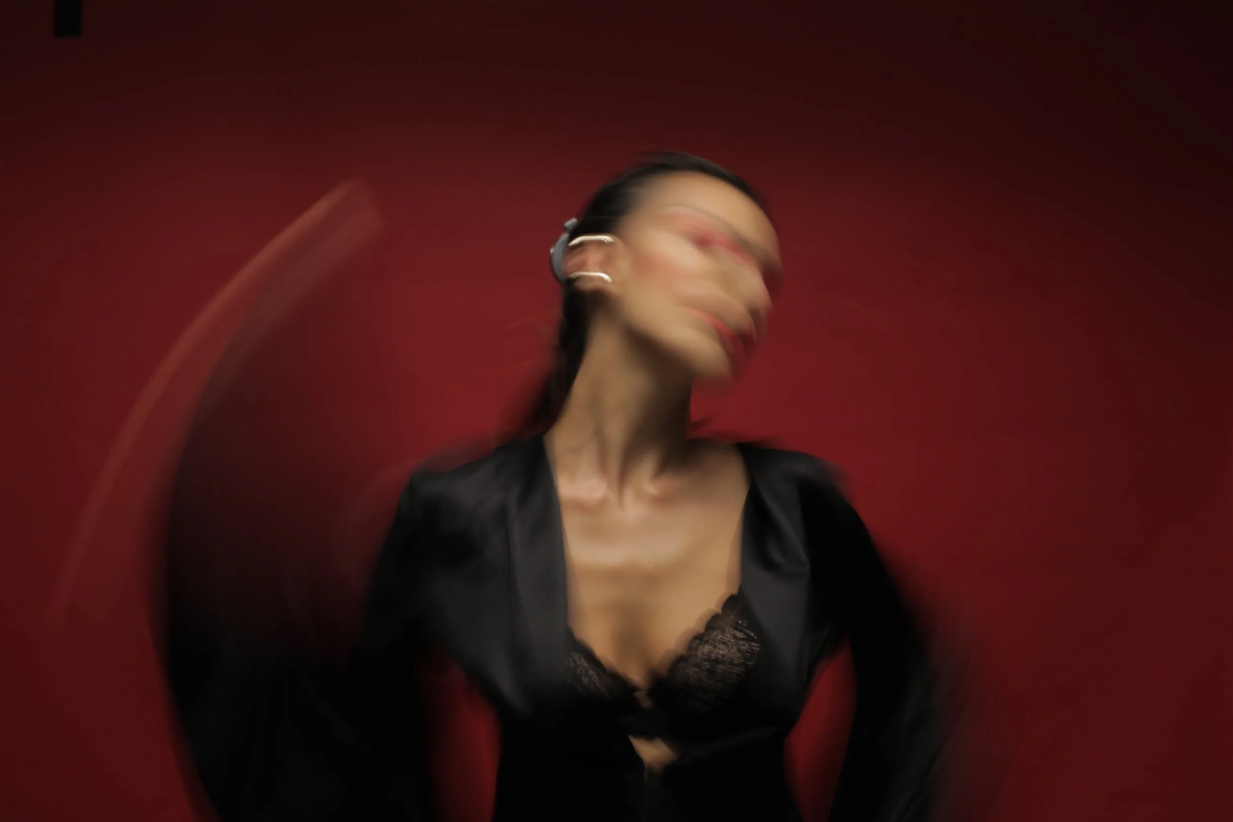 A woman with dark hair and glasses is smiling and wearing a black lace top. The photo has a motion blur effect, creating a sense of movement, with a red background, photographed by tim mattia