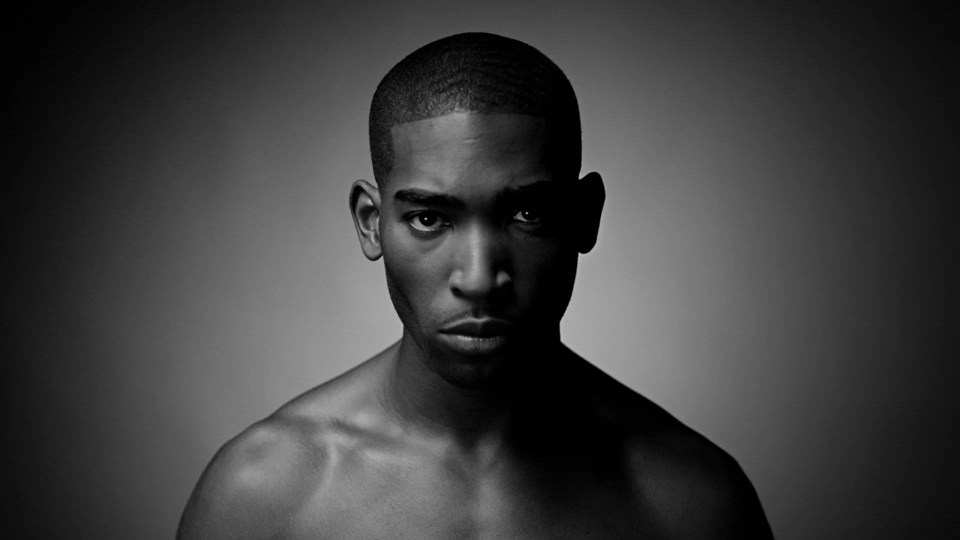 Black and white portrait of a young man with serious expression, looking directly at the camera, with short hair and bare shoulders.