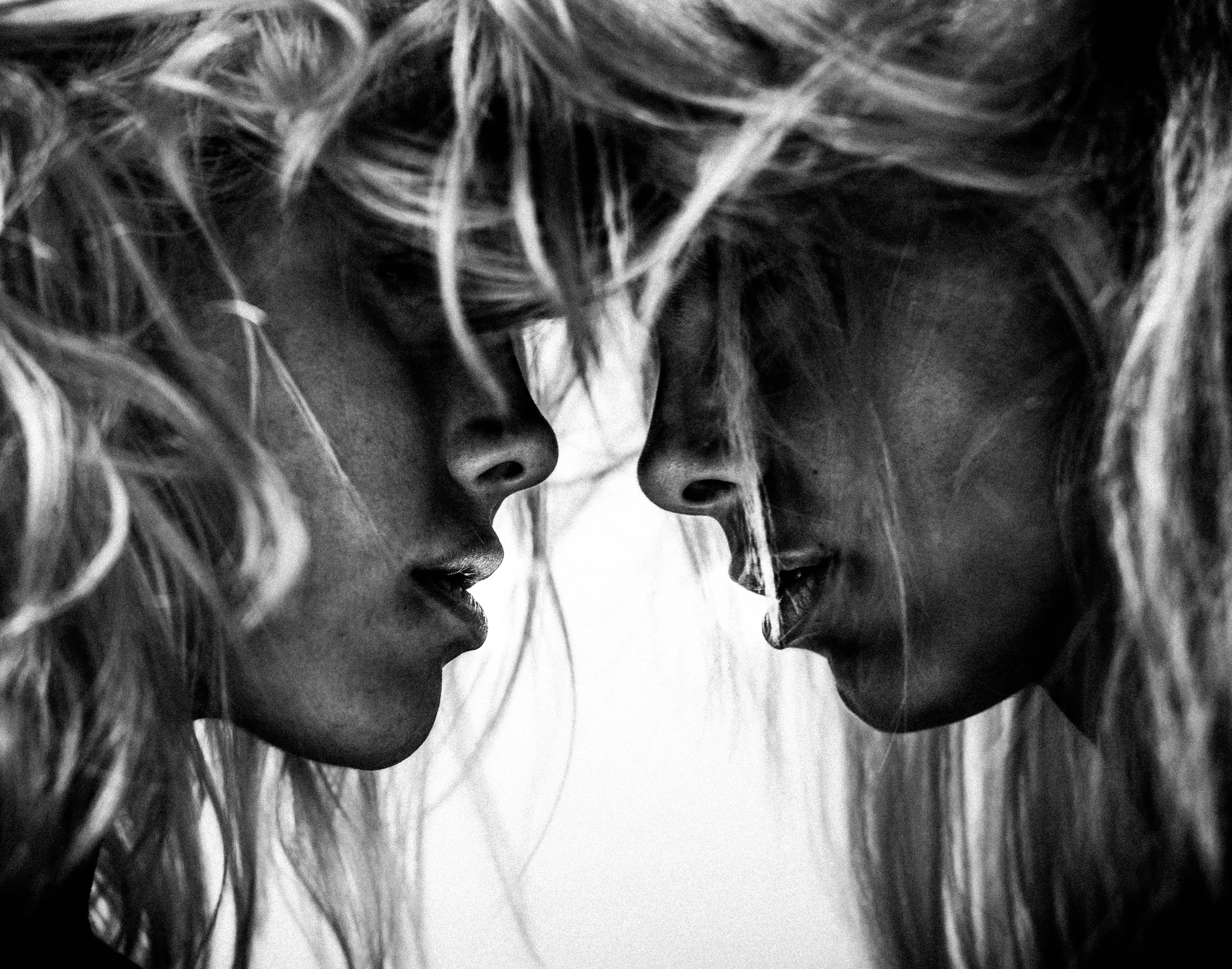 Close-up black and white photo of two young women with long, curly hair, noses, and foreheads touching.