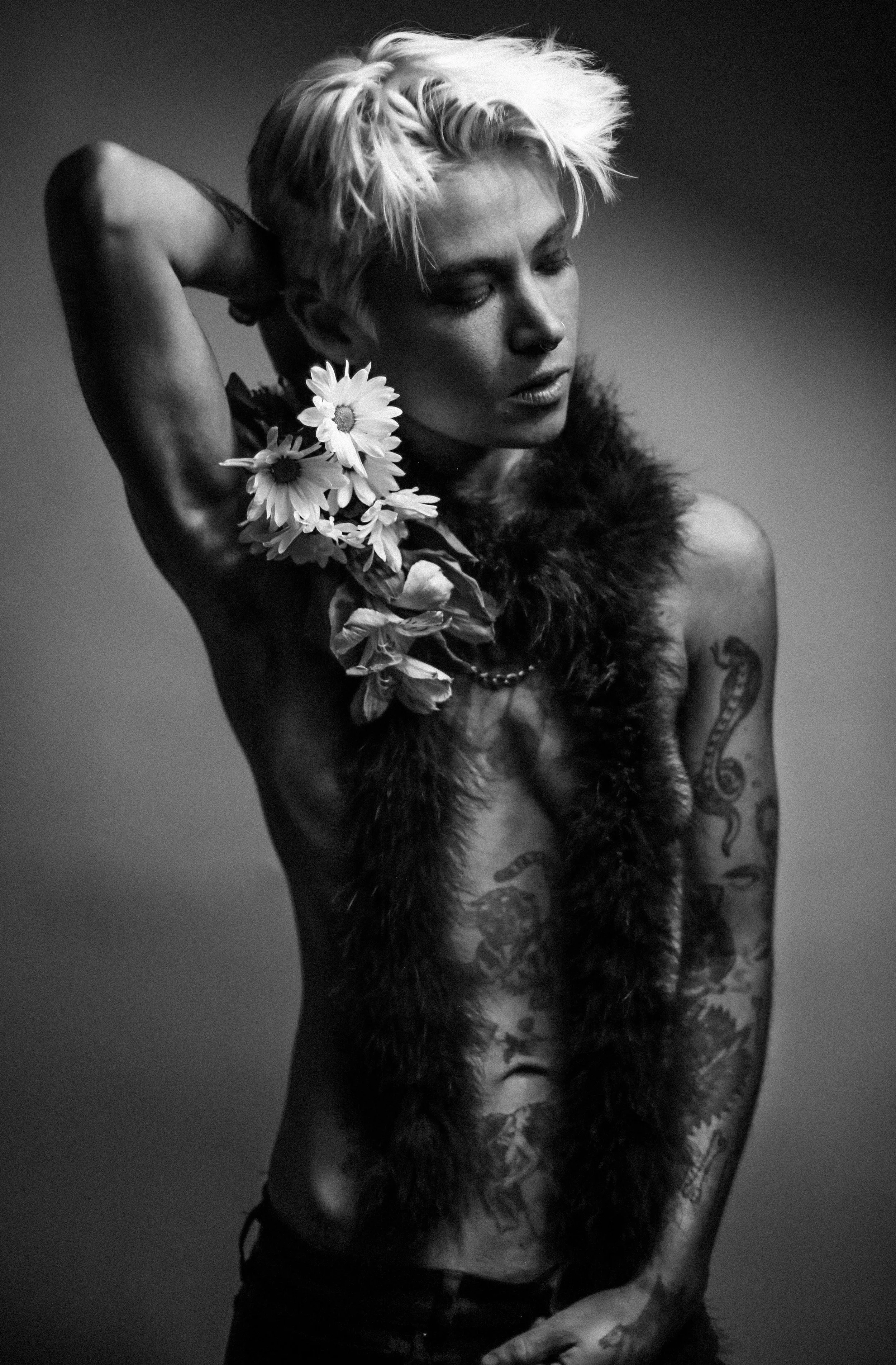 Black and white photo of a tattooed woman with short blonde hair, holding her arm behind her head, wearing a fur scarf, with daisies near her neck.