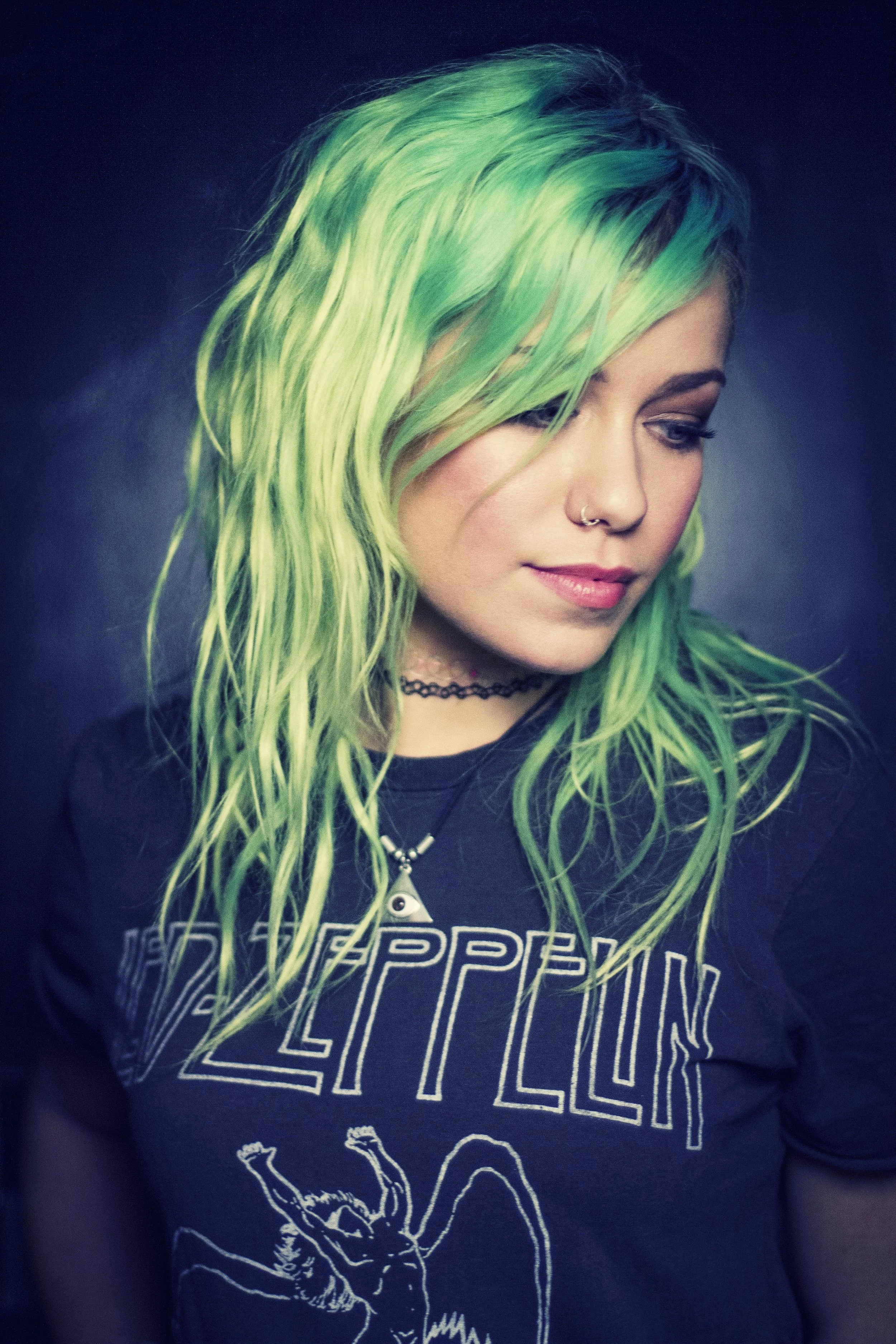 A woman with long, wavy, green hair, wearing a black Led Zeppelin t-shirt, a black choker, and a nose ring. She has a subtle makeup and a necklace with a pendant. The background is dark.