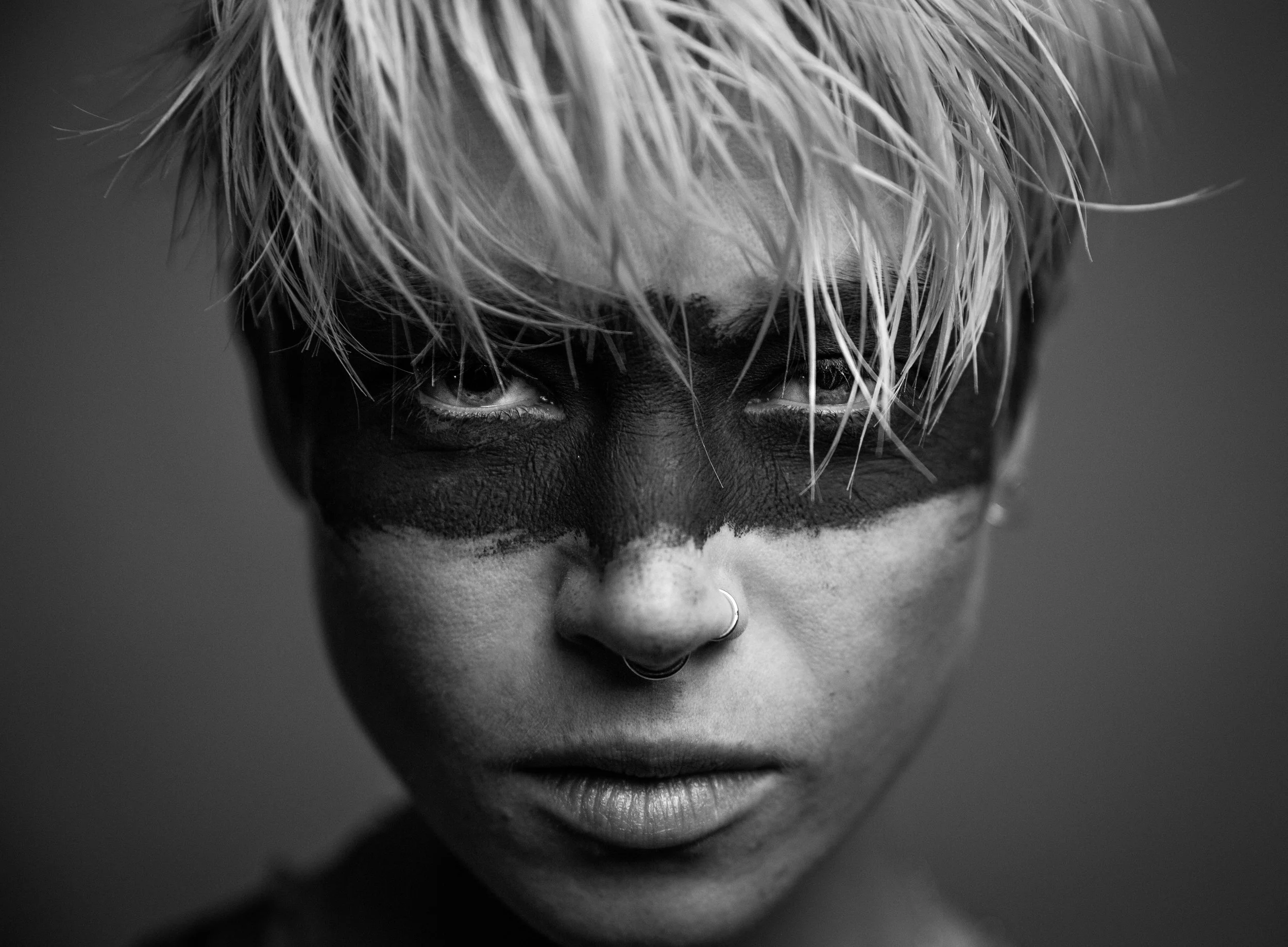 Close-up black and white portrait of a person with bleach blonde hair covering their eyes, with visible nose, lips, and a nose ring.