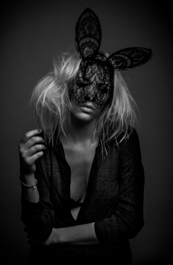 A woman with messy blonde hair wearing a lace bunny mask, dressed in a dark, open shirt, posed against a dark background in black and white, photographed by tim mattia