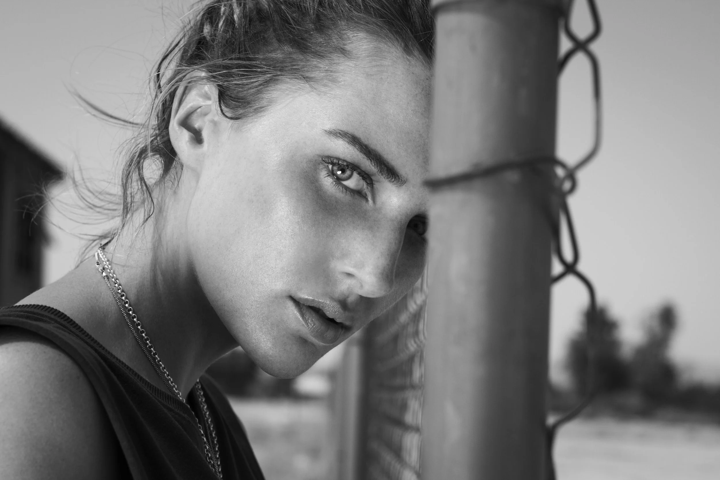 A black-and-white close-up of a woman's face with her hair tied back, wearing a chain necklace, with a focused expression, partially obscured by a chain-link fence.