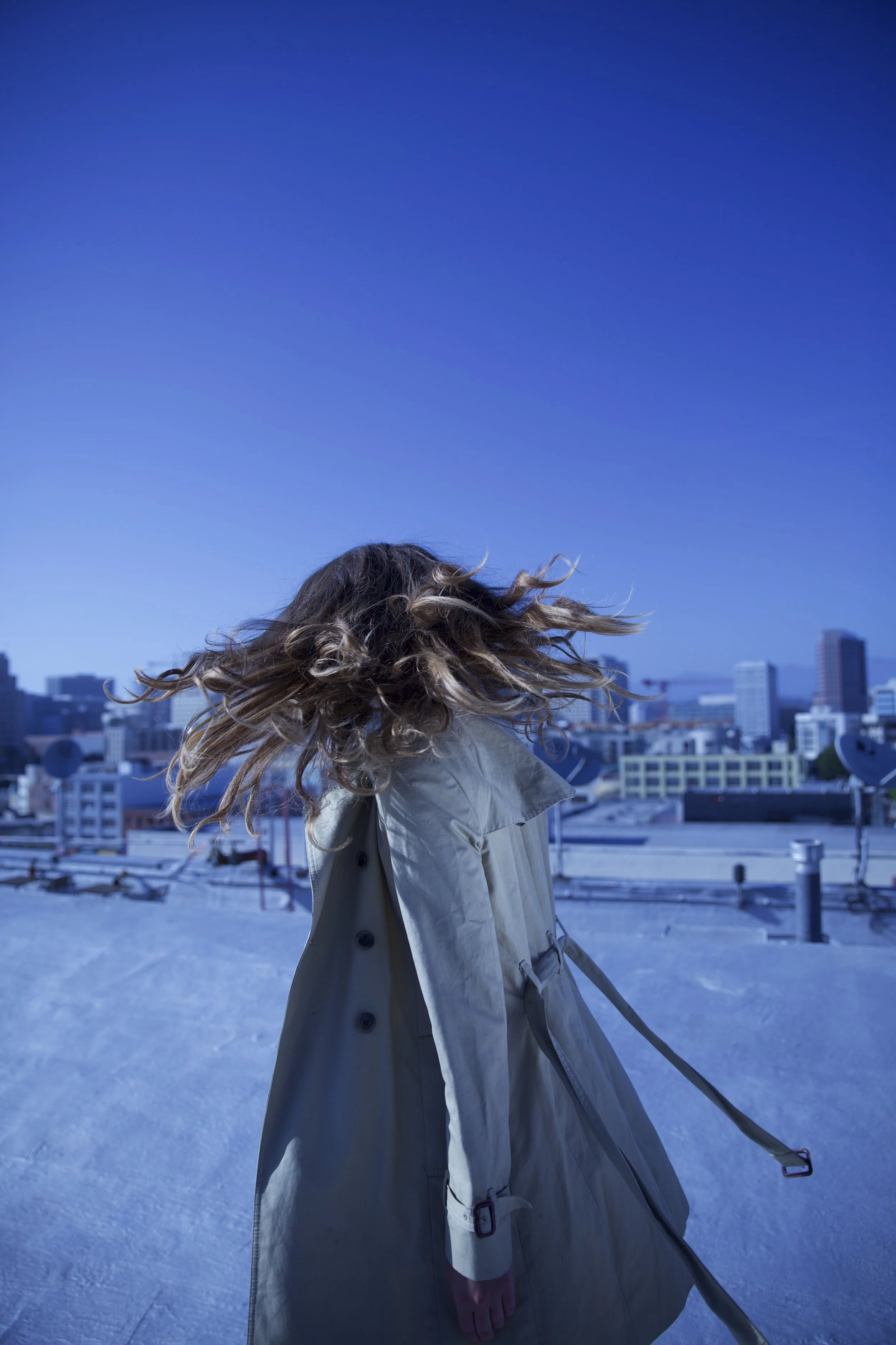 Person with curly hair blowing in the wind, wearing a beige trench coat, holding a leash, on a rooftop with city buildings in the background under a clear blue sky.