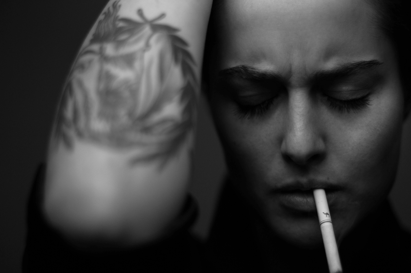 A close-up black and white photo of a woman with closed eyes, holding her forearm with a tattoo of a leaf pattern, and a cigarette in her mouth.