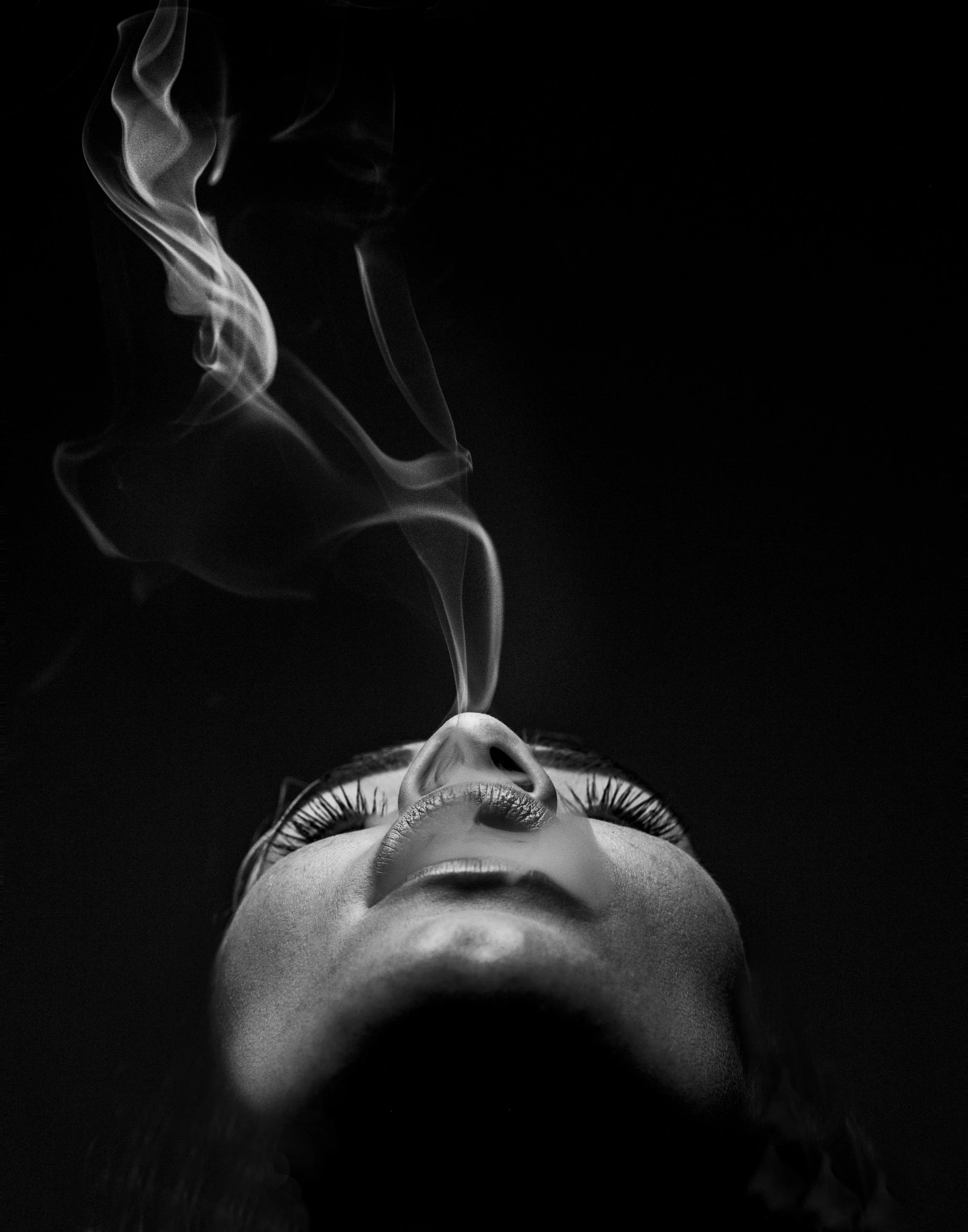 A black-and-white photo of a human face lying down on a dark surface, viewed from below. The person has eyelashes and closed eyes, with smoke rising above their face.