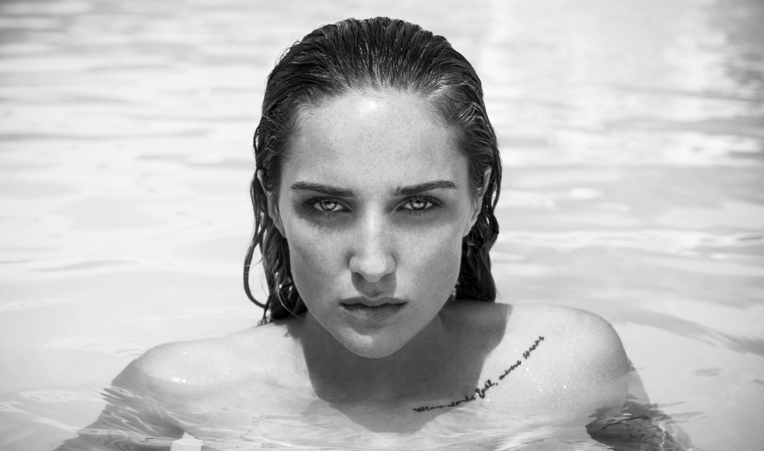 Black and white photo of a woman partially submerged in water, with her head and shoulders visible. She has wet hair slicked back and intense eyes staring at the camera, with water droplets on her face and shoulders.