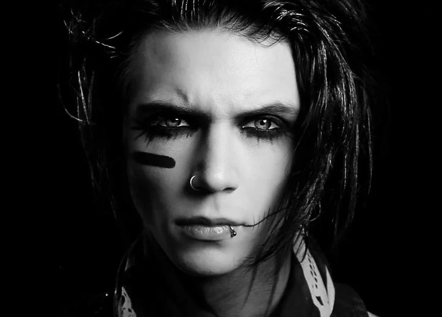 Close-up black and white portrait of a person with intense facial expression, black lipstick, facial piercings, dark eye makeup, and hair styled in a messy, alternative fashion.