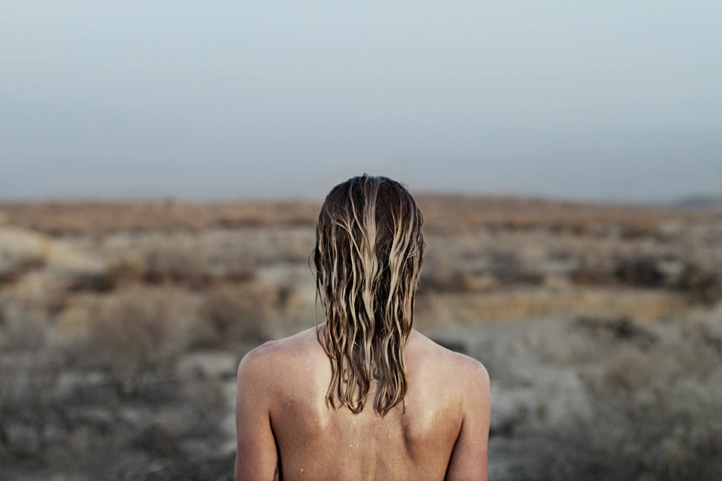 A person with long wet hair standing on a rocky beach with the ocean and sky in the background.