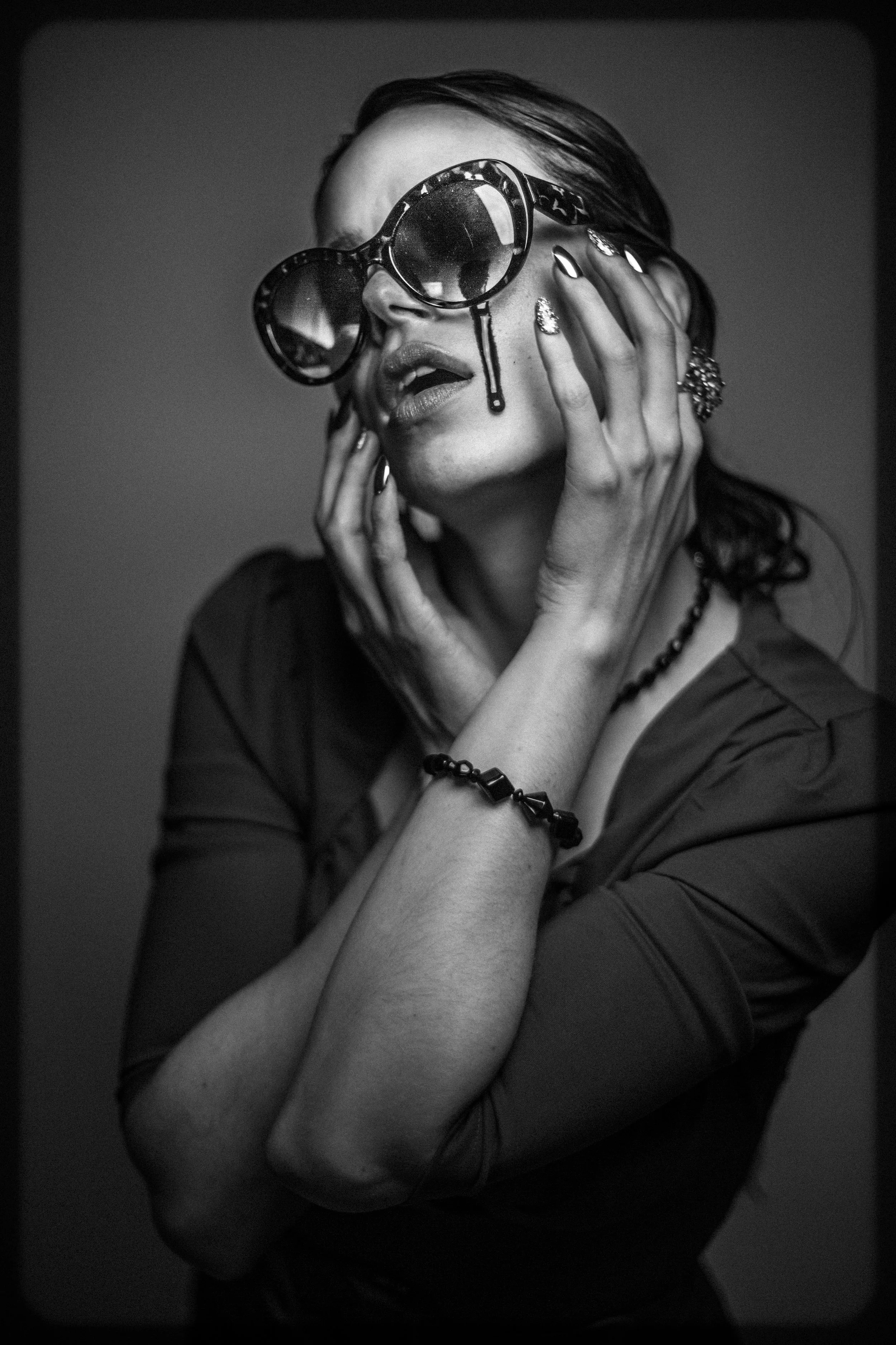 A woman wearing large sunglasses, touching her face with her hands, with her mouth slightly open, in a black and white photo.