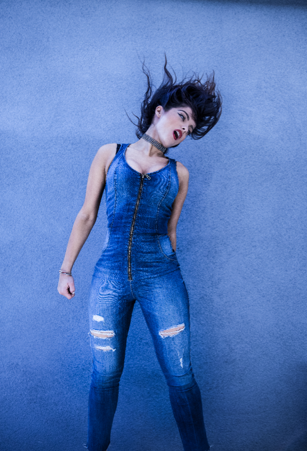 bebe rexha with dark hair in a blue denim jumpsuit with rips, standing against a blue wall, with hair flying and expressive pose photographed by tim mattia