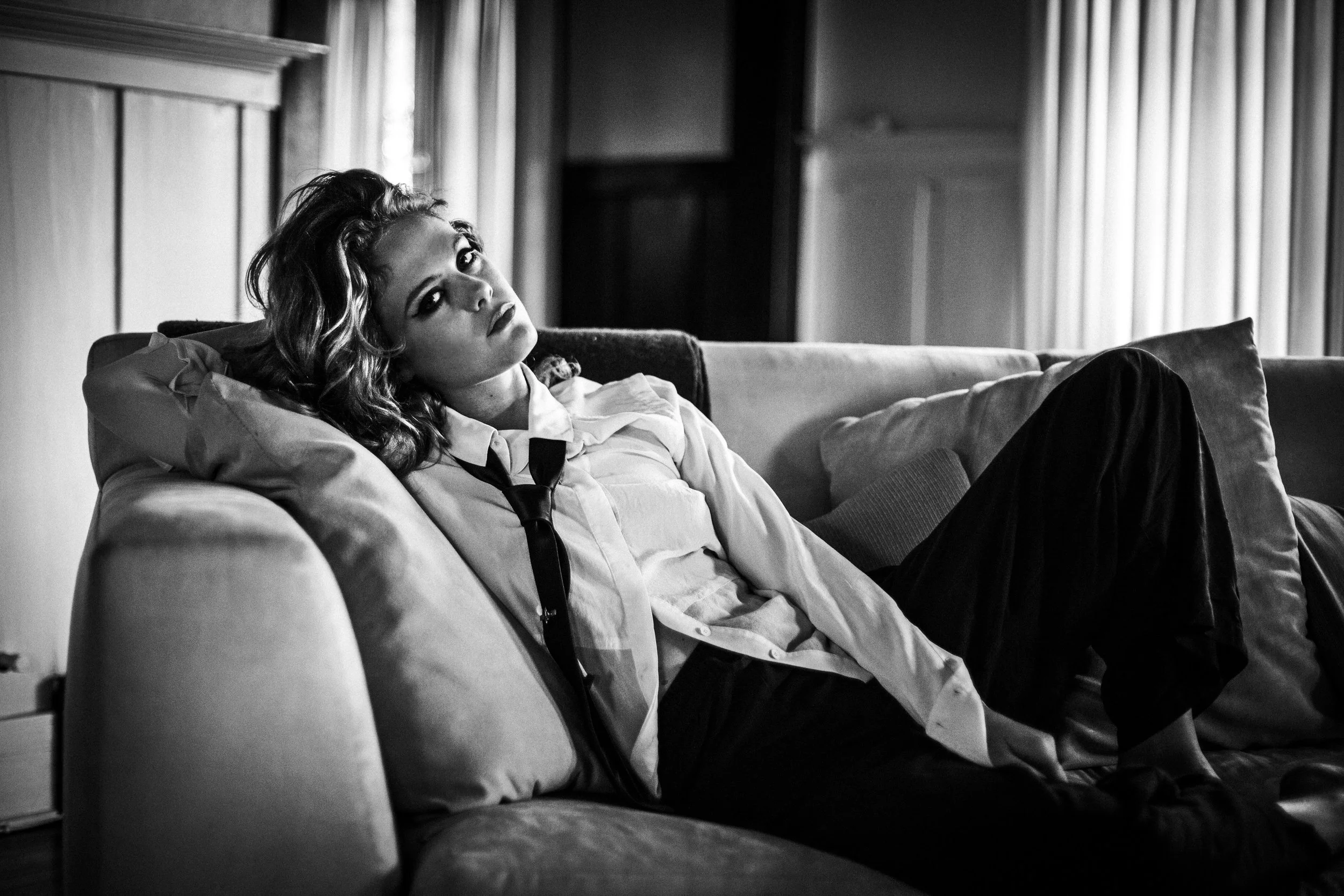 A woman with long, curly hair wearing a white shirt and loosened tie, sitting on a sofa in a room with draped curtains, gazing thoughtfully.