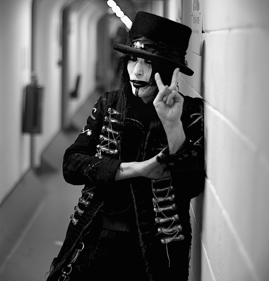 Black and white photo of Tommy the guitarist with Motley Crue leaning against a wall in a hallway backstage at wembley, making a peace sign with their hand, photographed by tim mattia