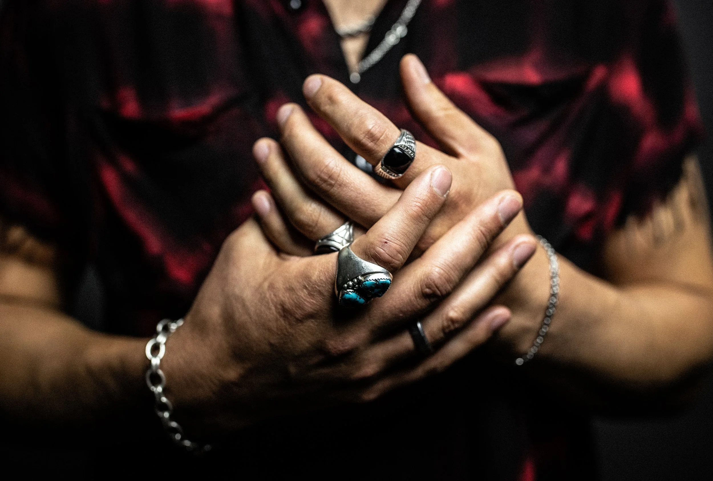 Close-up of a person wearing multiple silver rings and a silver bracelet, with hands placed on chest, against a black and red patterned shirt.