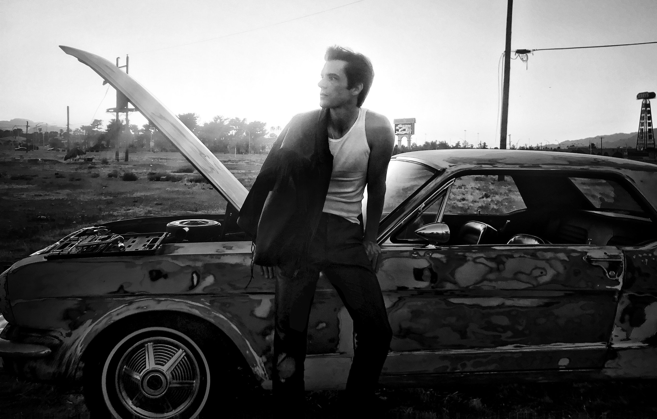 A black and white photo of a Brandon Flowers lead singer of The Killers leaning against a vintage car with the hood open, standing outdoors during sunset, with power lines and distant hills in the background, photographed by tim mattia