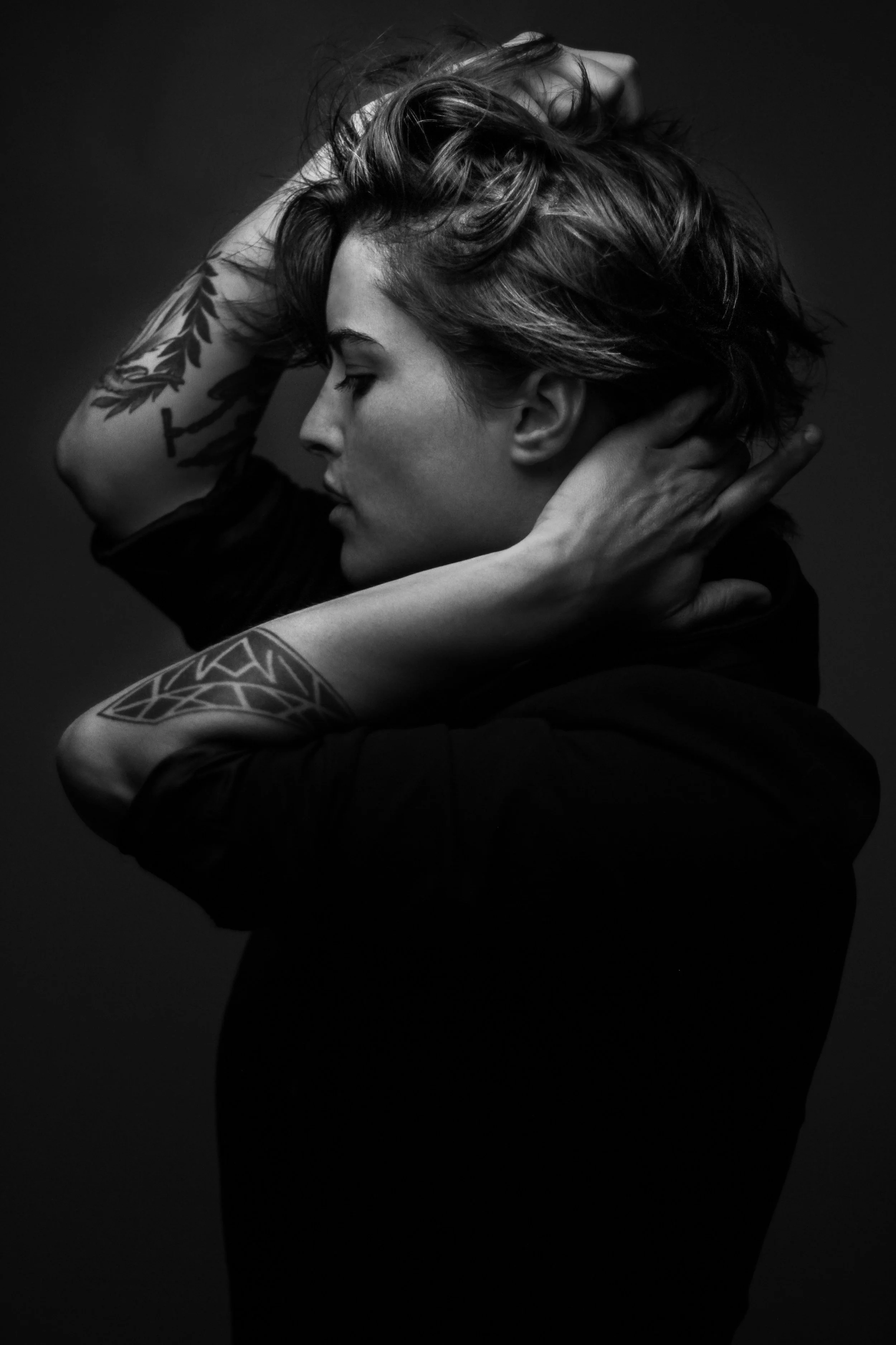 Black-and-white photo of a woman with short, tousled hair, eyes closed, head tilted back slightly, with one hand behind her neck and the other on her head, showcasing tattoos on her arms.
