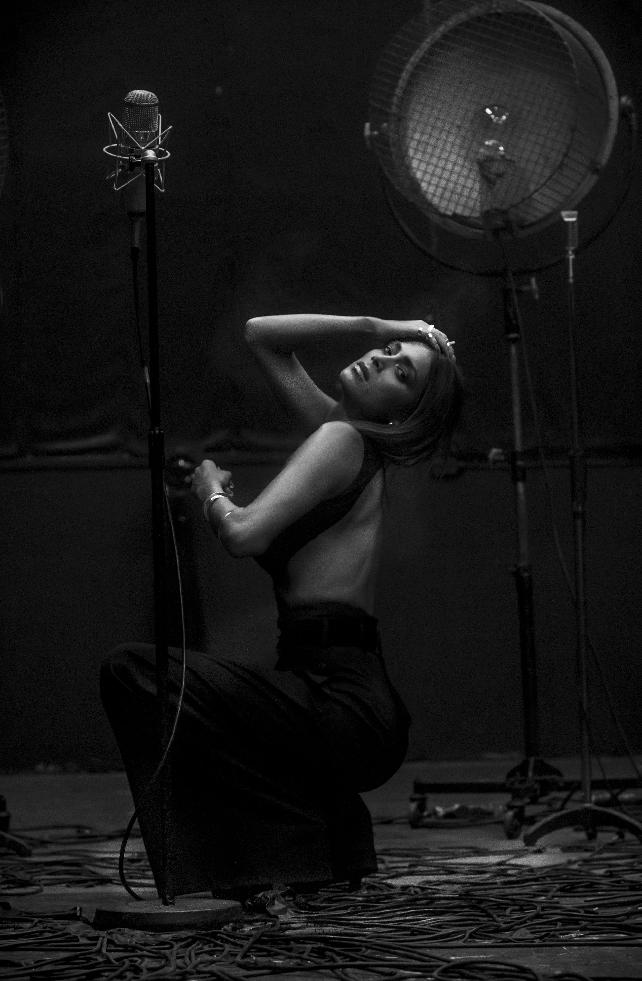 A woman with long hair and a sleeveless top, kneeling on the floor, looking at the camera, surrounded by photography equipment, including lighting and cables.