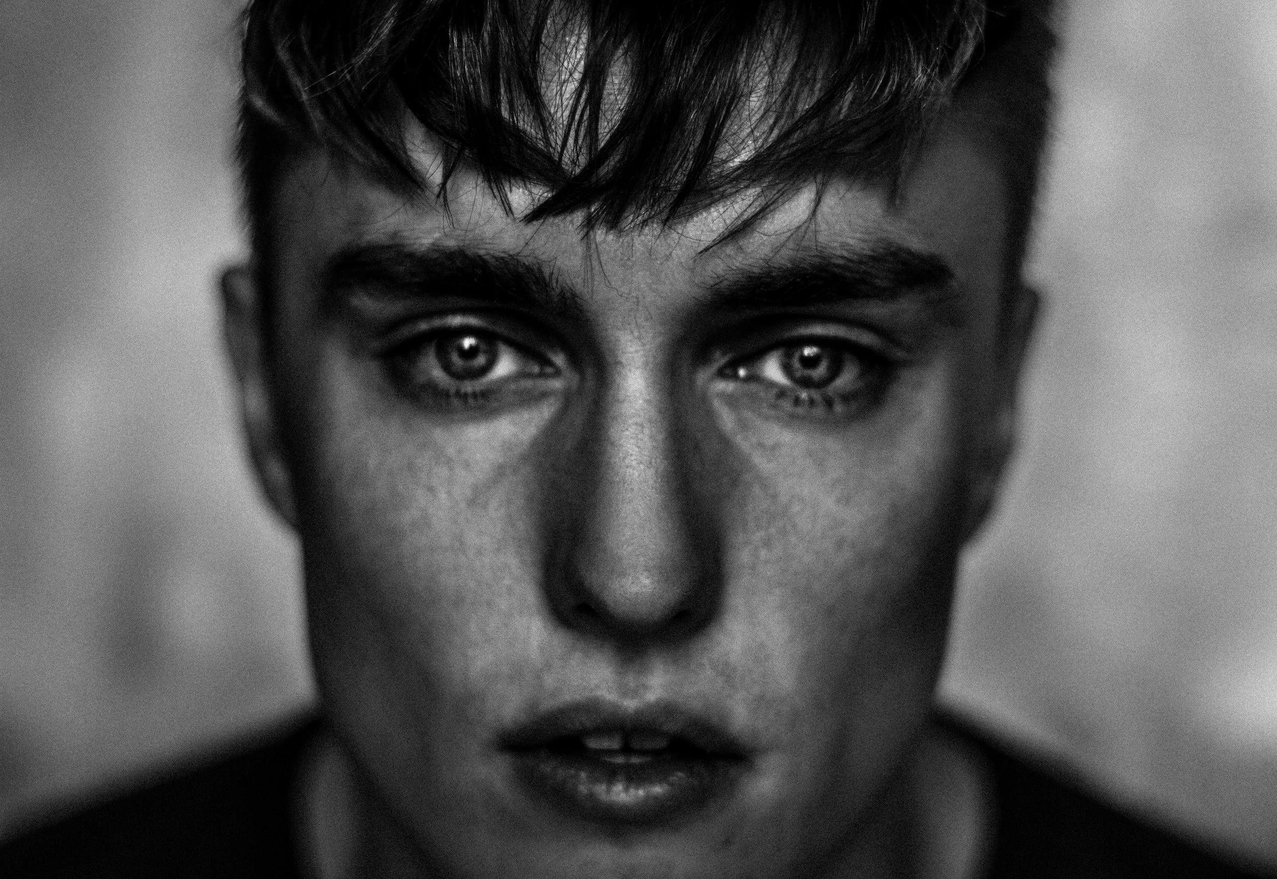 A close-up black and white portrait of a young man with intense eyes, messy hair, and a serious expression.