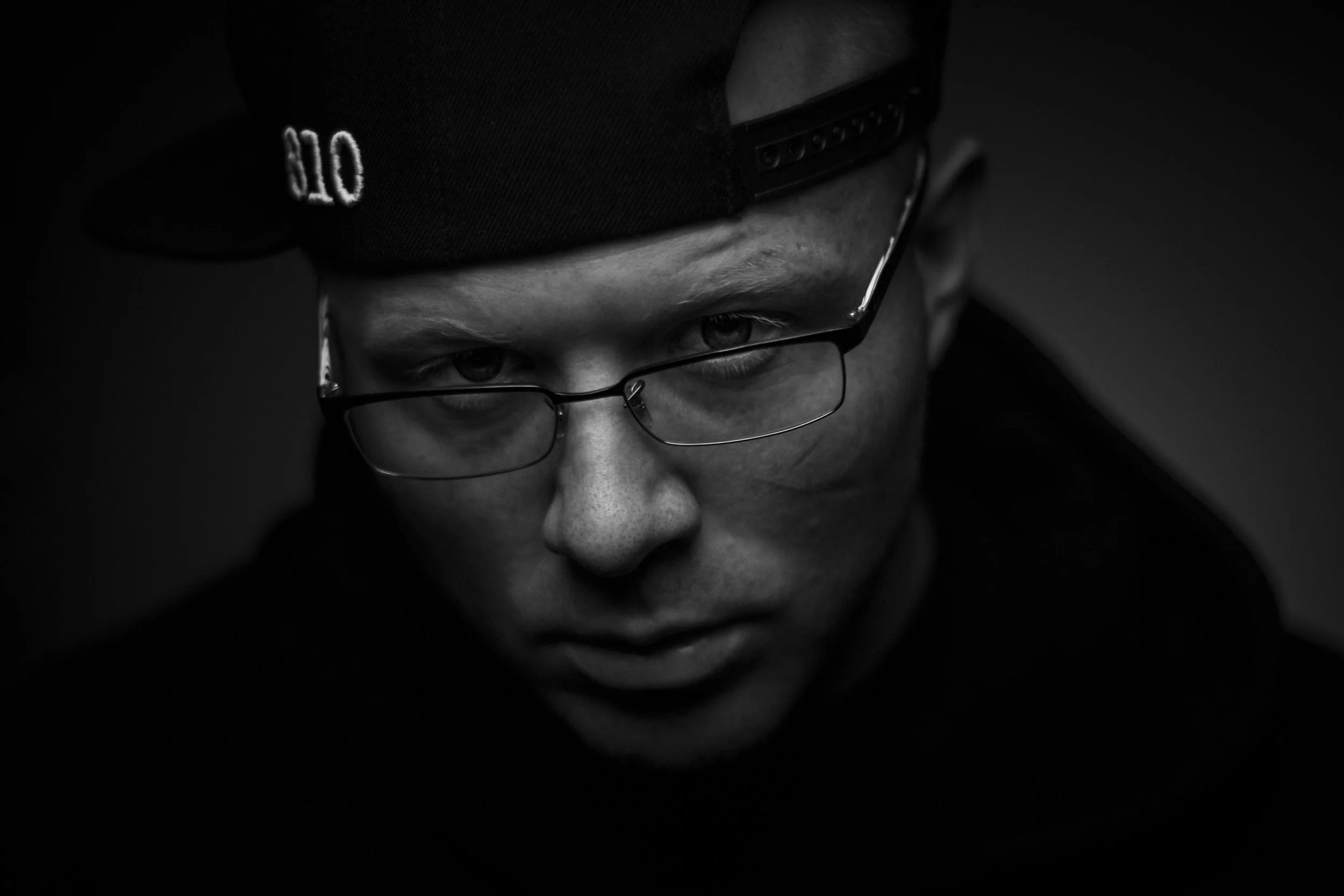 Close-up black and white photo of a man wearing glasses and a backward baseball cap, looking down with a serious expression.
