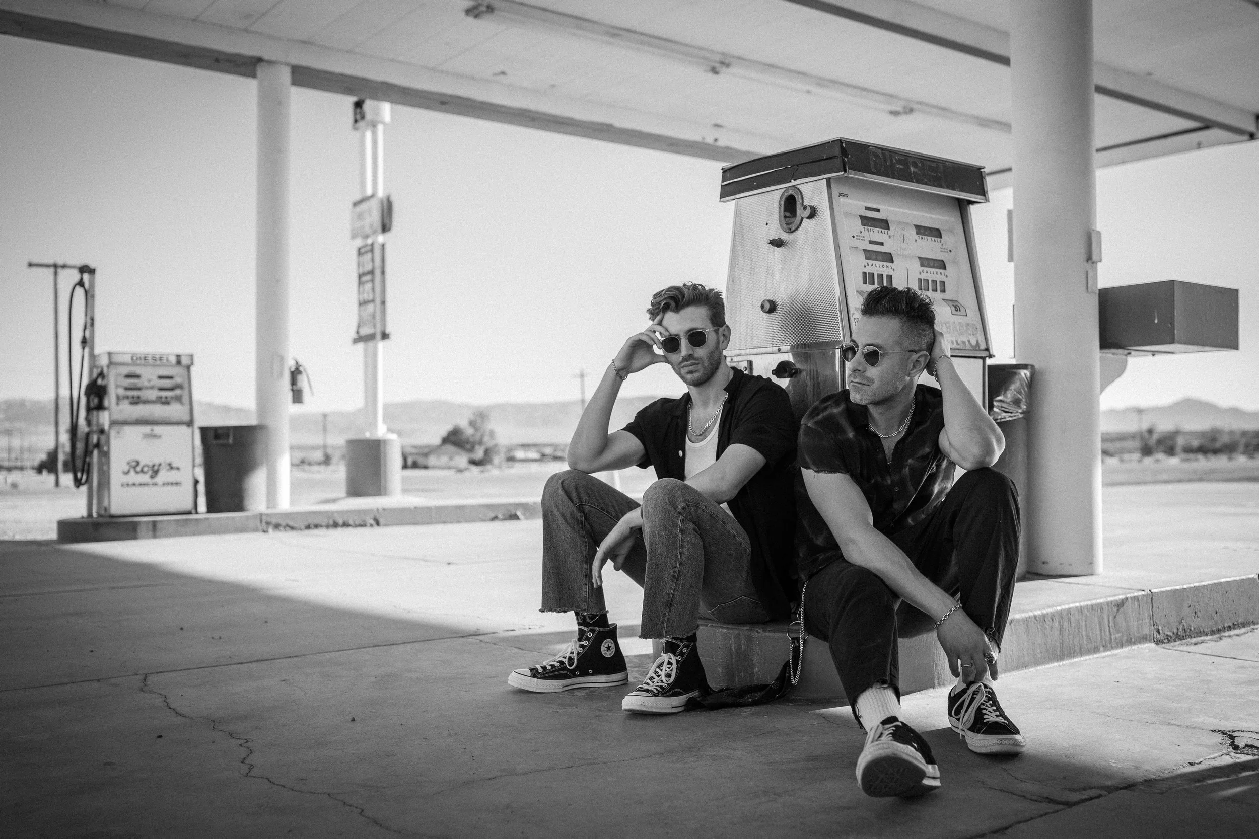Two young men with sunglasses sitting on the ground at a gas station, one adjusting his glasses and the other looking away, with gas pumps and rural landscape in the background.