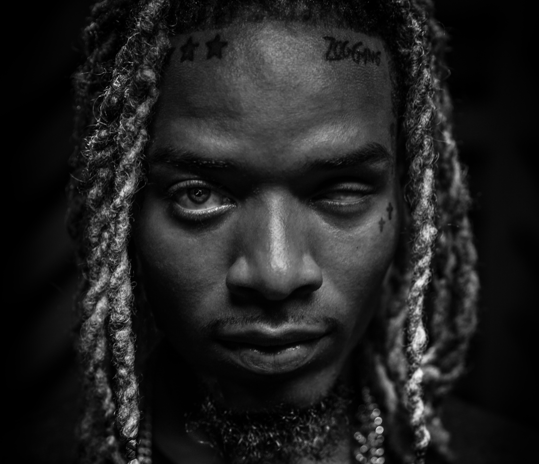 Close-up black and white portrait of a person with dreadlocks, tattoos on forehead and face, and an intense expression.