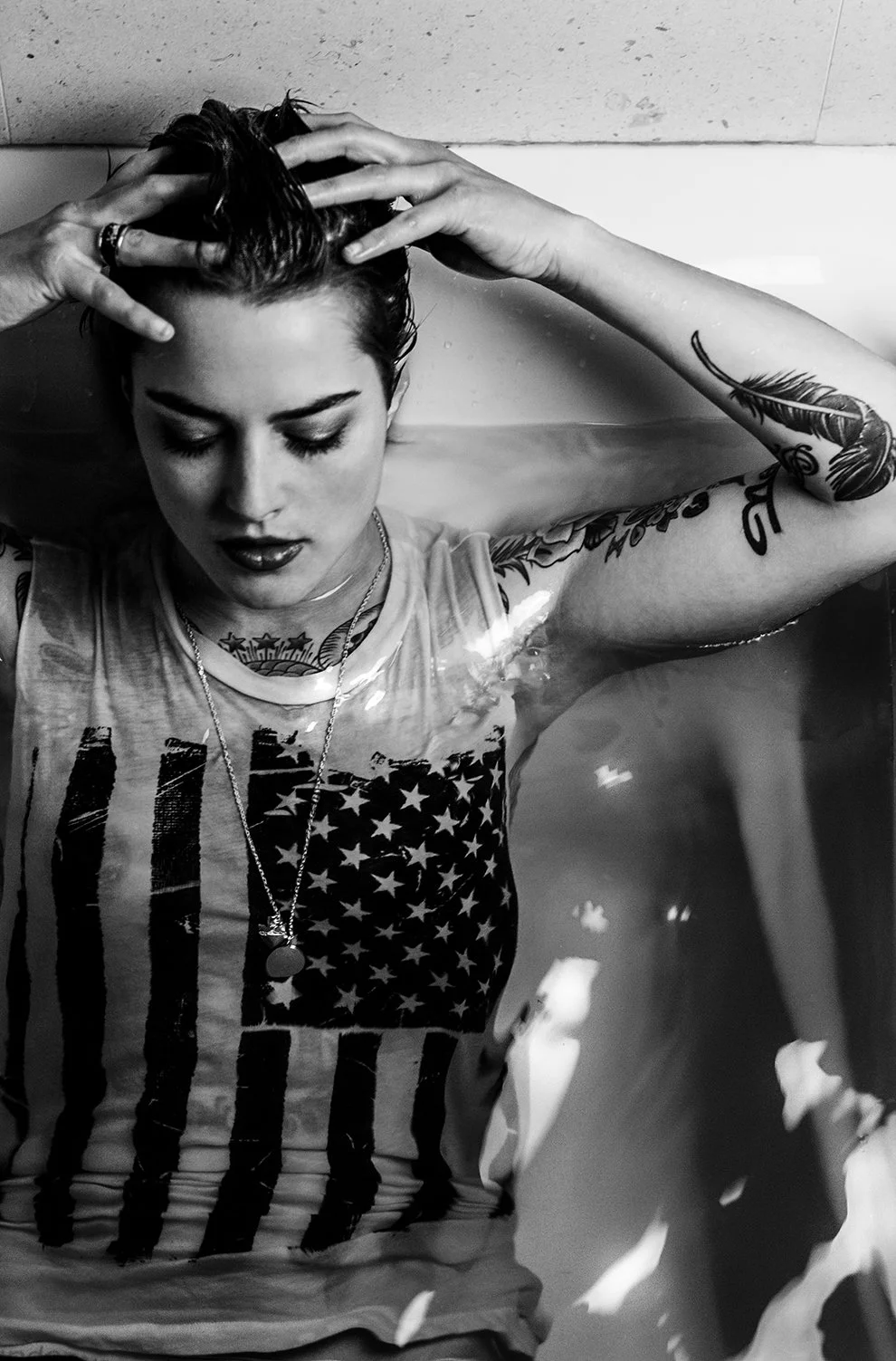 Young woman with tattoos on arms and chest, wearing an American flag tank top, submerged in water, with her hands in her wet, slicked-back hair, eyes closed, and a neutral expression.
