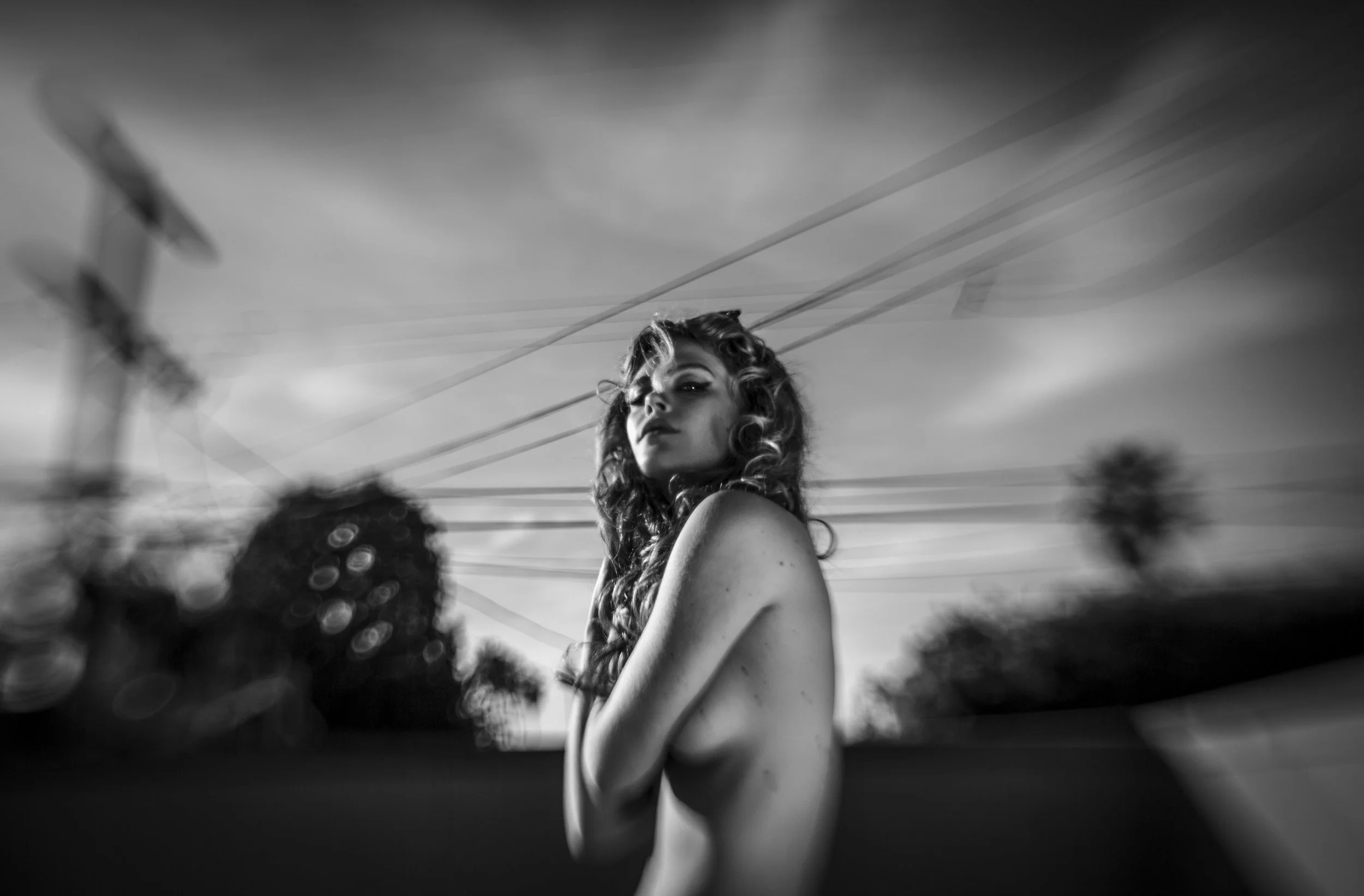 Black and white photo of a model and actor Chelsea Schuchman with curly hair looking over her shoulder, with out-of-focus objects in the foreground and cables or wires in the background photographed by tim mattia