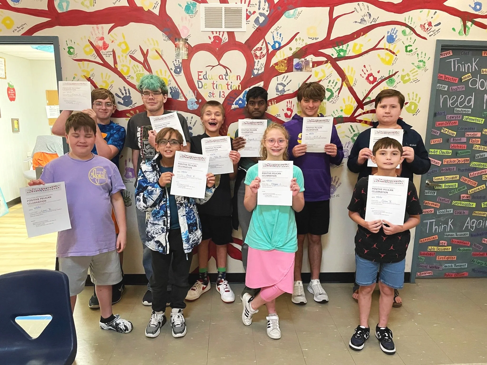 A group of young students indoors, holding certificates and posing in front of a decorated wall with colorful handprints and the words 'Education Definition' in the center. Some students are smiling, and they appear to have just received awards or recognition.