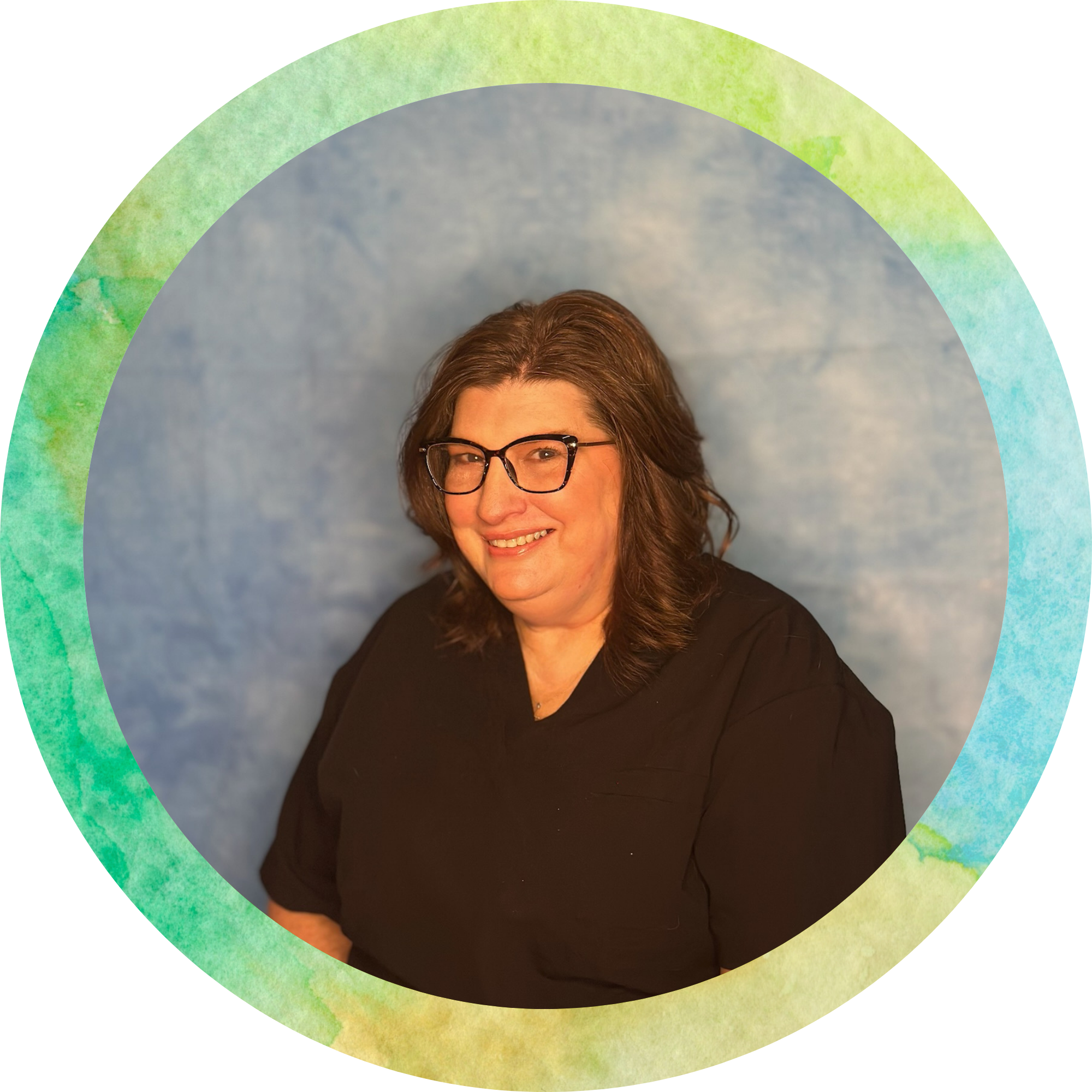 A woman with glasses smiling, wearing a black top, standing in front of a tie-dye background with shades of blue, green, and yellow.