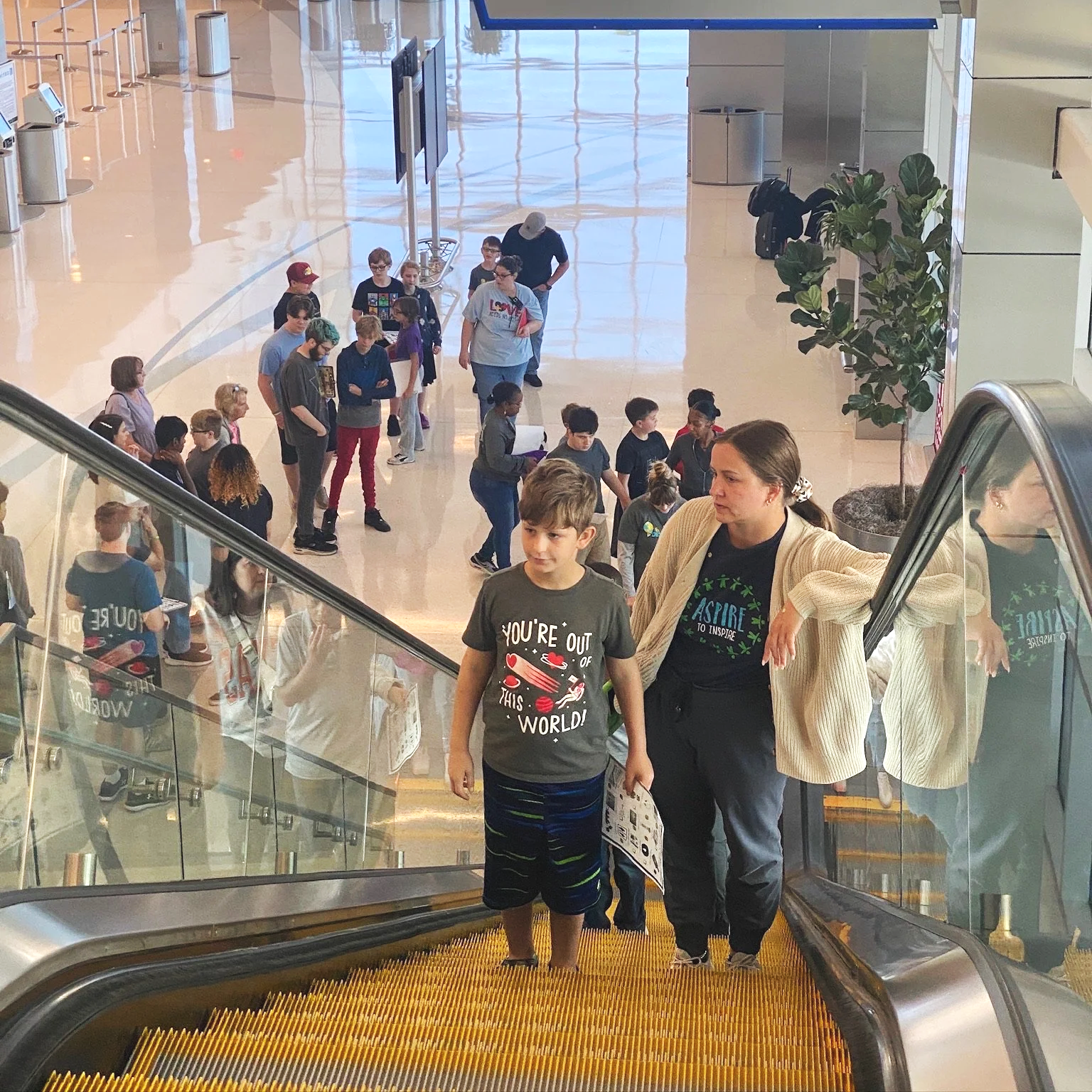 People ascending an escalator in an indoor public space, with some groups waiting in line and others walking. The area is well-lit with a polished floor and large windows.