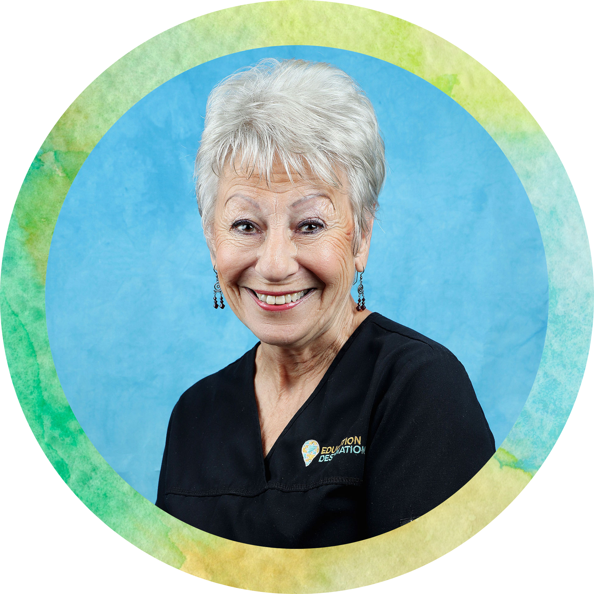 Close-up of an elderly woman's face smiling, with short gray hair and earrings, wearing a black shirt with a 'Education' patch, set against a vibrant, colorful background.